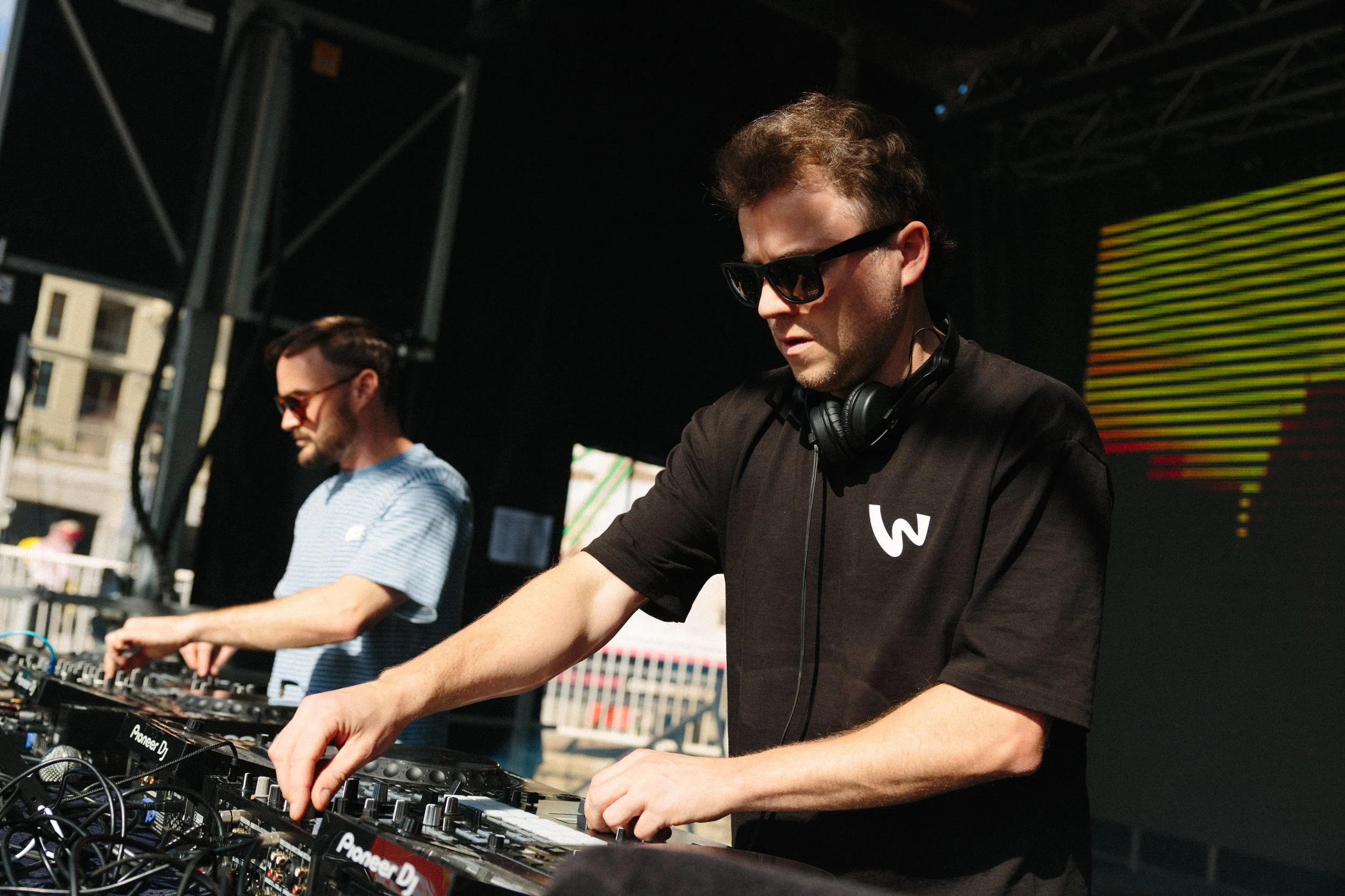 Two men wearing sunglasses and headphones are DJing at an outdoor event with musical equipment, including a Pioneer DJ controller, in front of them at night.