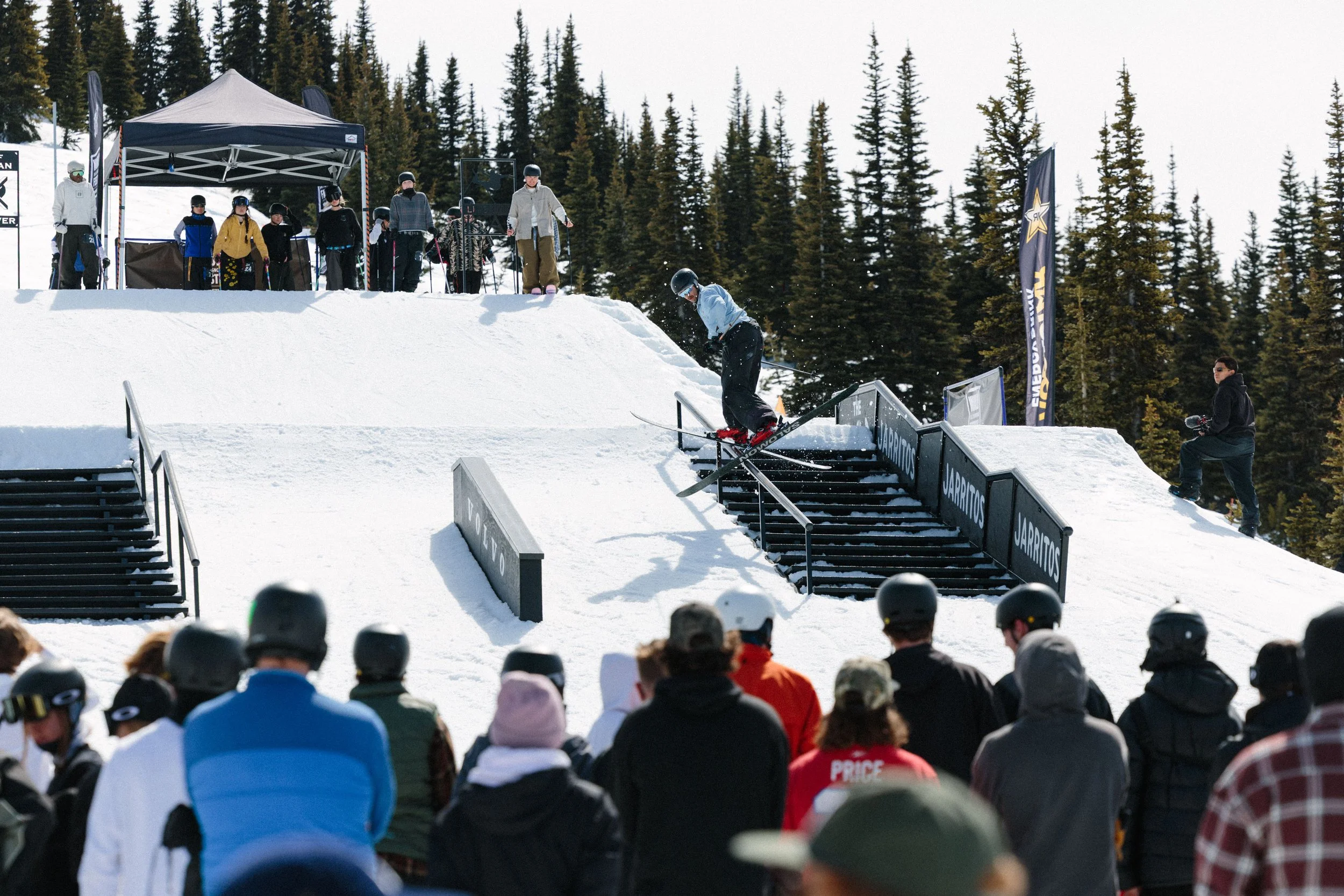 A skier is jumping off a staircase rail at a snowboarding event, with a crowd of spectators watching in the foreground and a group of people, including other skiers and organizers, on the snow behind a tent. Tall pine trees are in the background.
