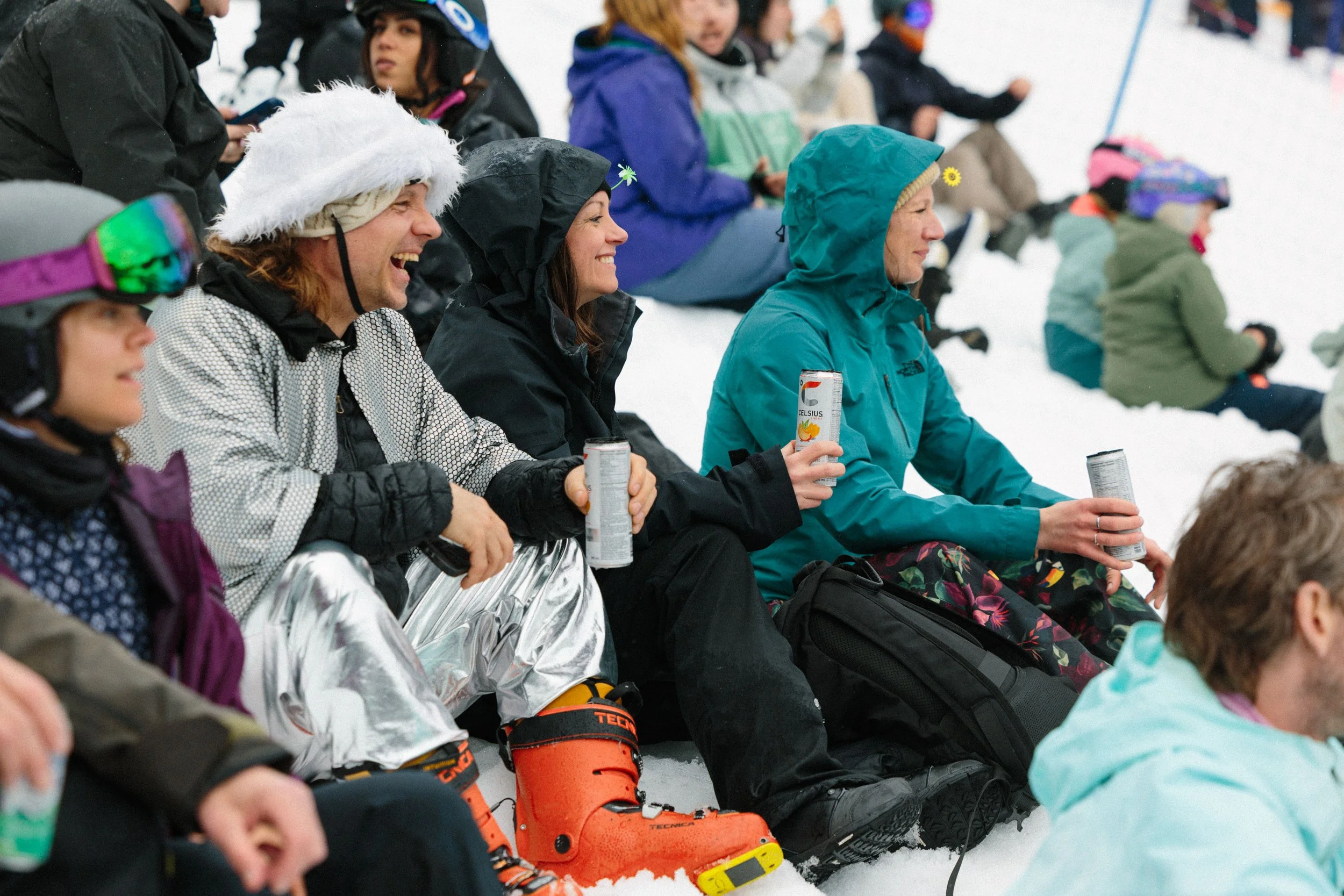 Group of people sitting in the snow, dressed in winter clothing, enjoying a winter outdoor event. Some are holding cans of drinks, and some are wearing ski boots and goggles.