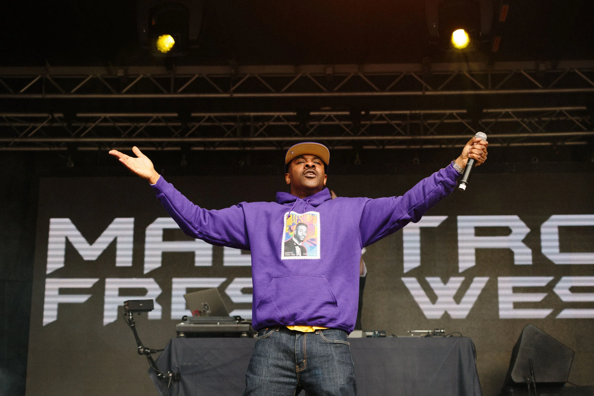 A man in a purple hoodie and a tan baseball cap standing on a stage with arms outstretched, holding a microphone in his right hand. There is stage equipment and a large LED screen behind him with some large text partially visible.