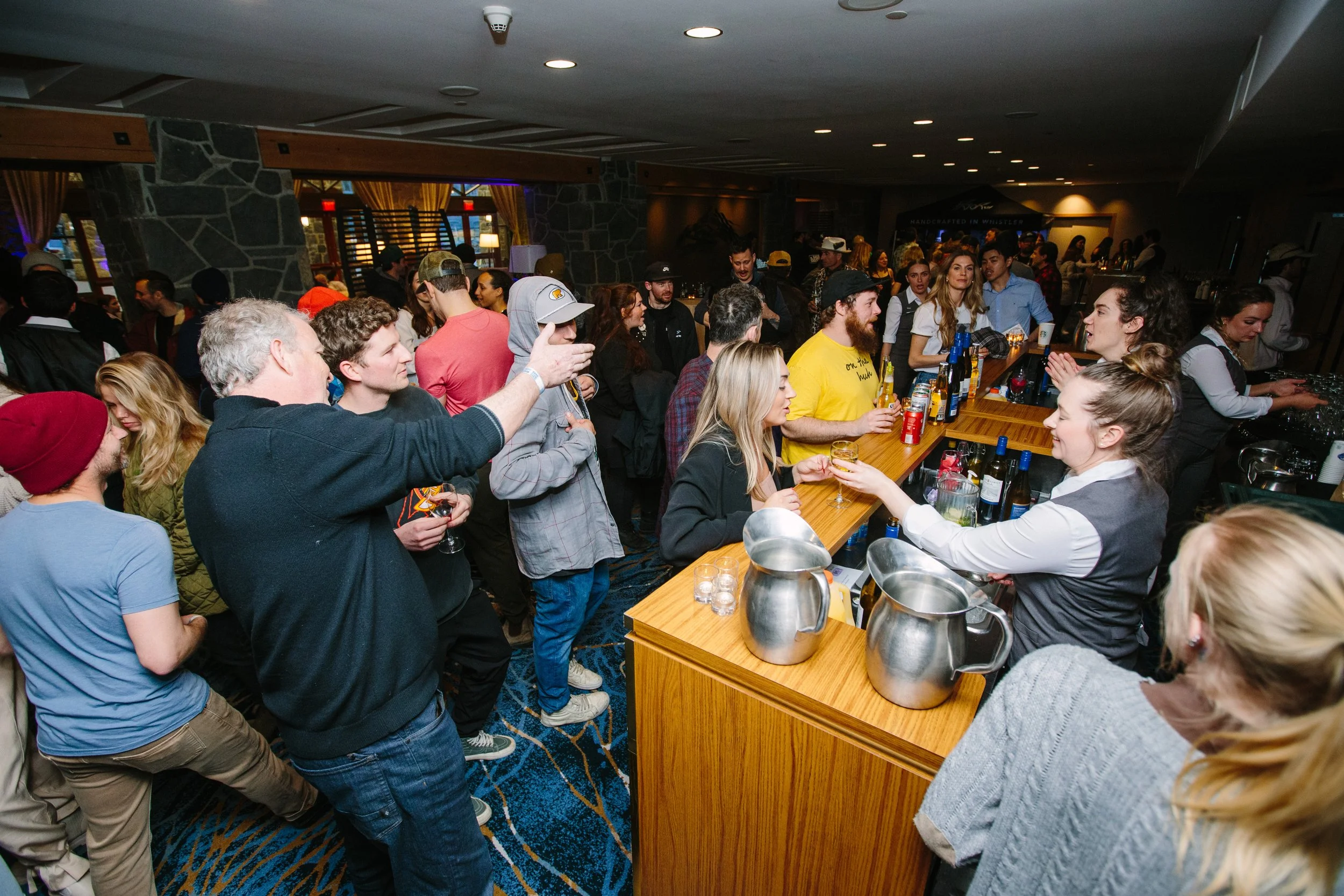 People gathered at a bar in a busy indoor setting, some ordering drinks, others talking and socializing.