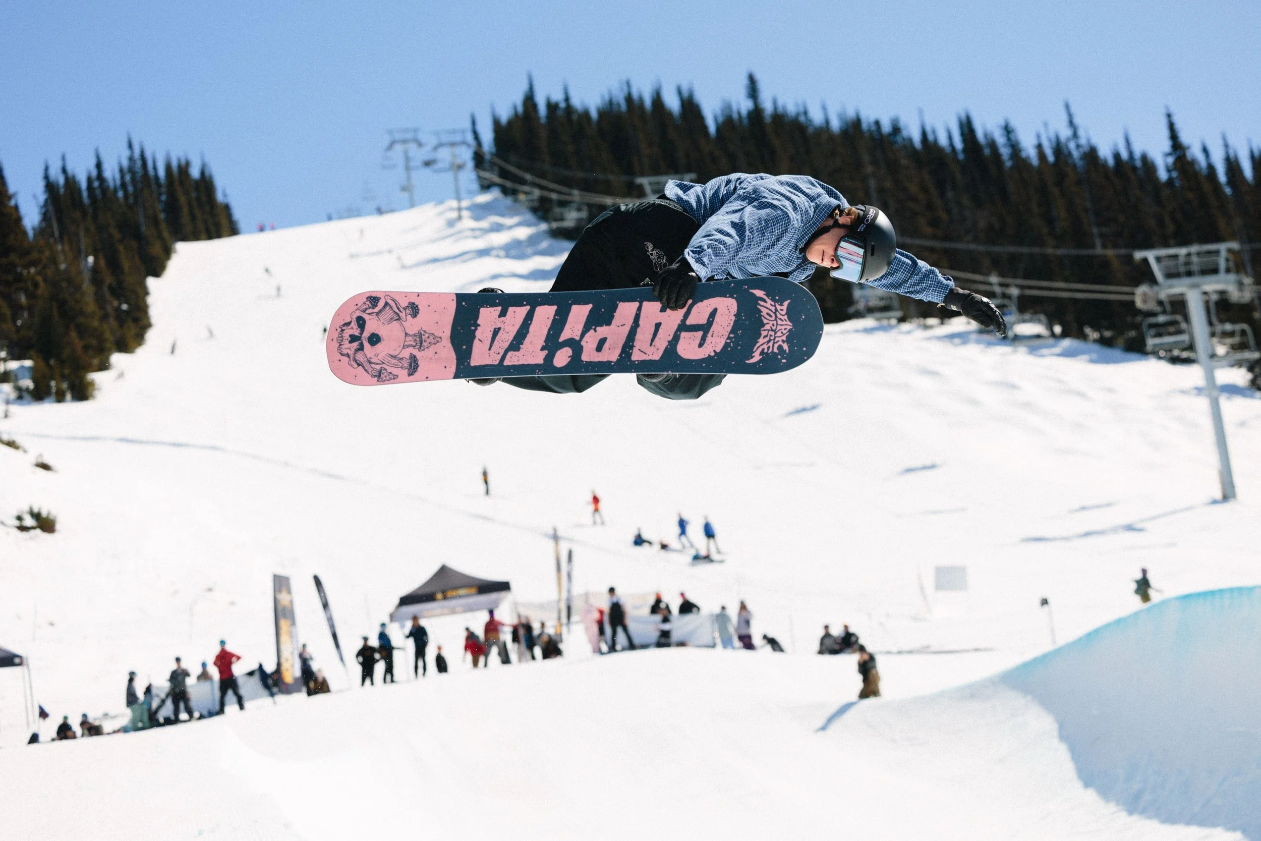 A skier in a blue checkered jacket, black helmet, and black gloves is mid-air performing a jump on a snowy ski slope with a pink and purple snowboard. In the background, there are skiers and snowboarders, ski lifts, and snow-covered trees under a cle