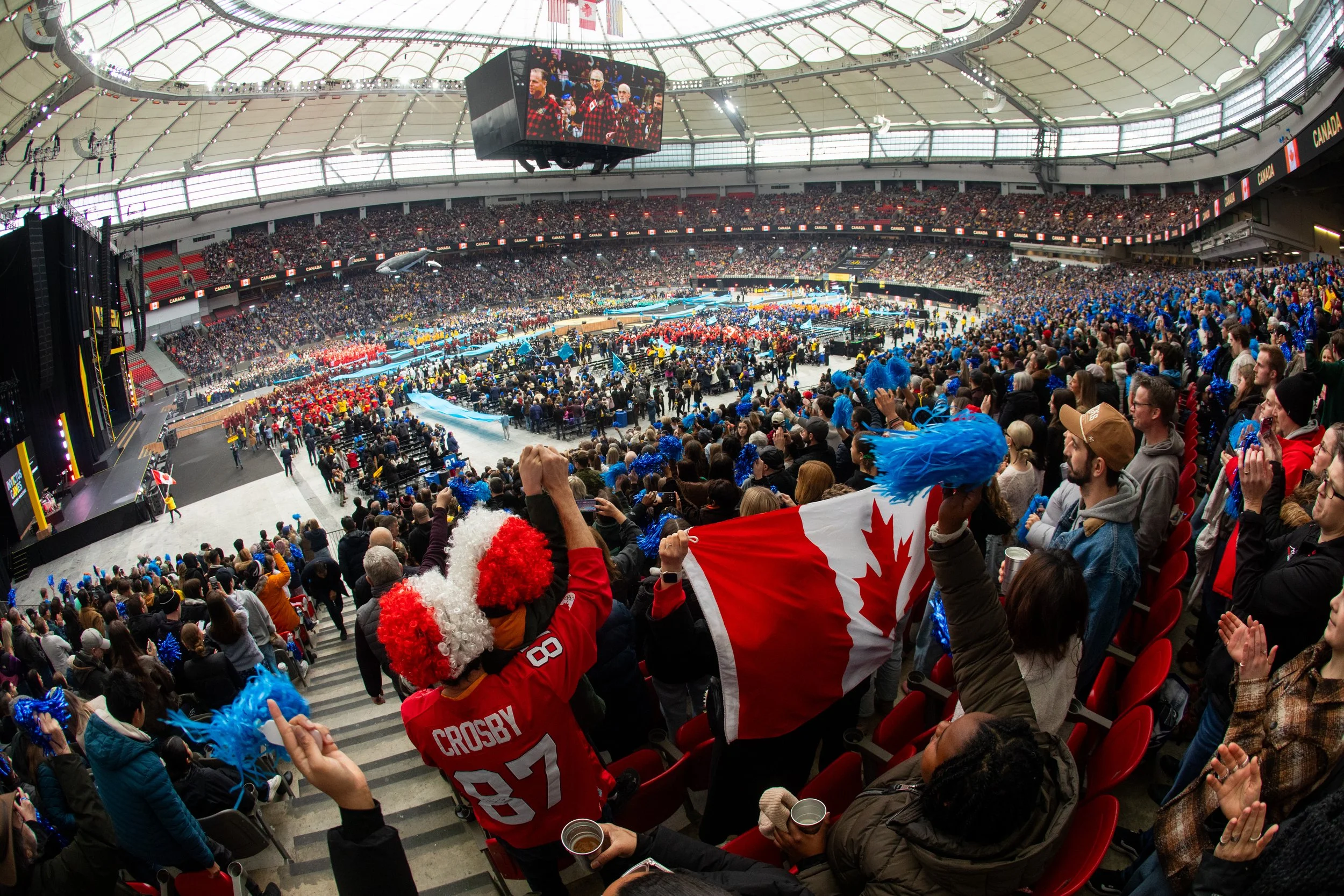 Crowd of spectators at a large indoor sports stadium watching a game or event, with some fans waving Canadian flags and dressed in team apparel.
