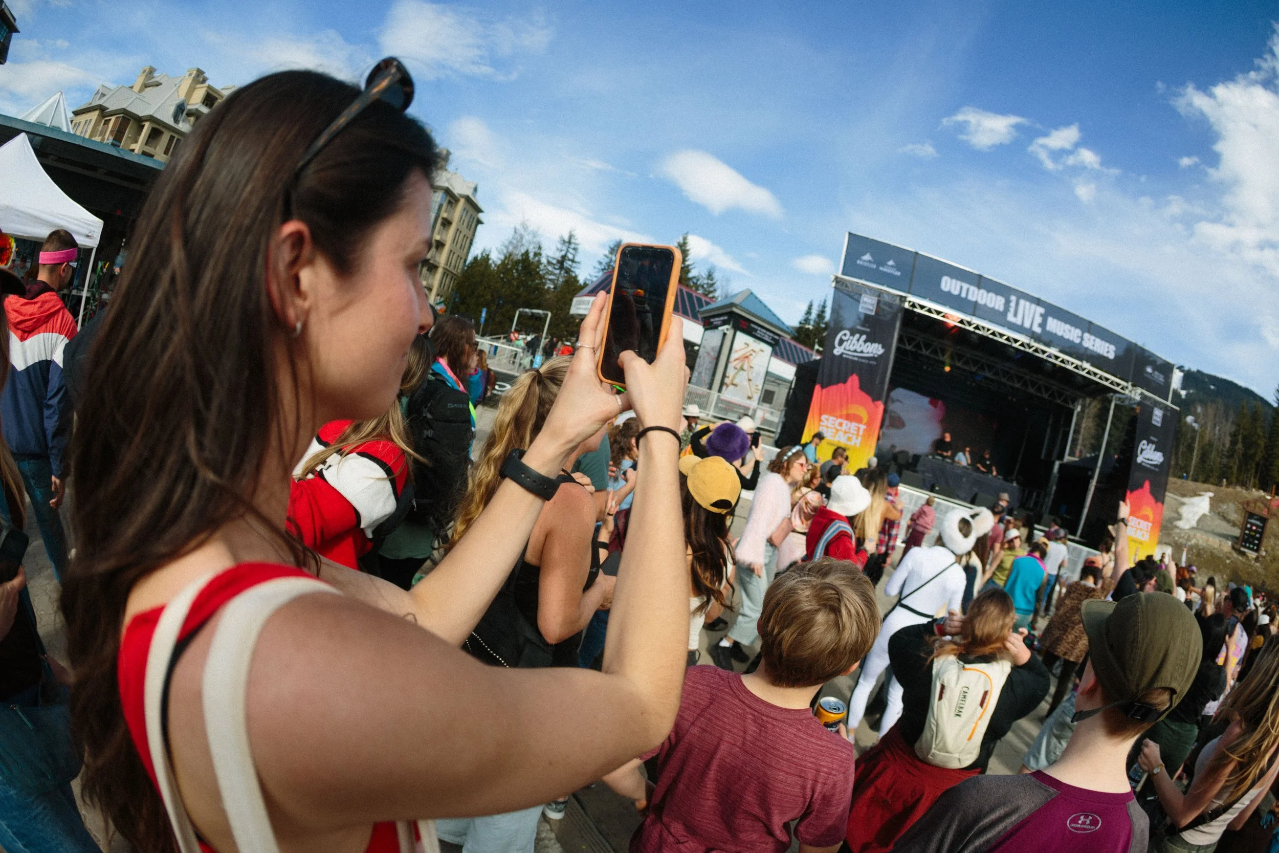 People gathered at an outdoor music festival, with a stage labeled "Outdoor Live Music Series" and the words "Secret Beach" visible on a banner. The audience is watching a performance under a partly cloudy sky.