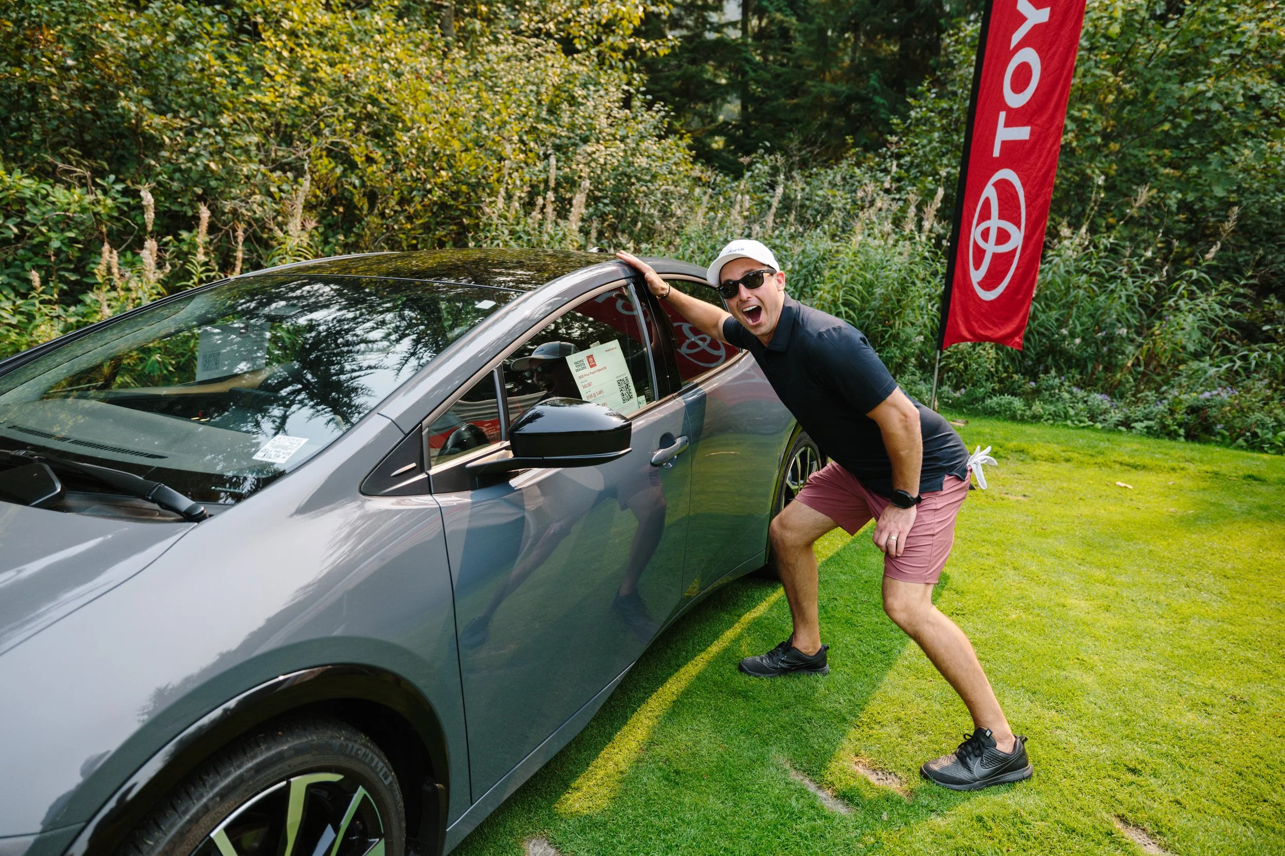 A man wearing sunglasses, a white cap, black shirt, and pink shorts, posing excitedly next to a gray sports car in a grassy area with a Toyota flag in the background.