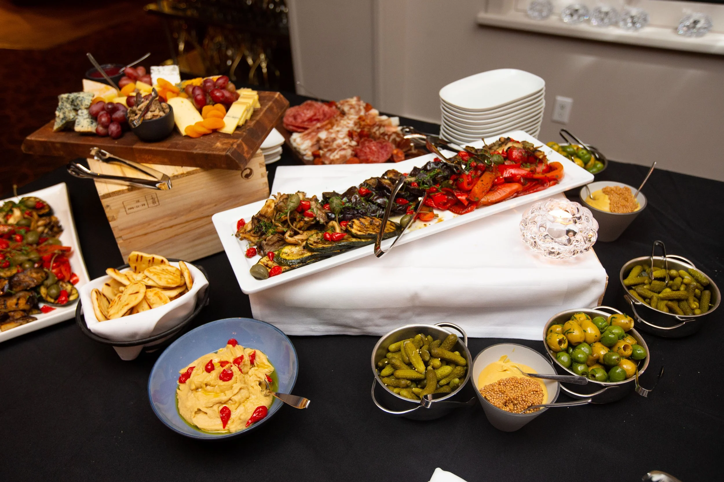Buffet table with assorted cheeses, grapes, dried apricots, charcuterie, roasted vegetables, hummus, pita bread, pickles, and olives.