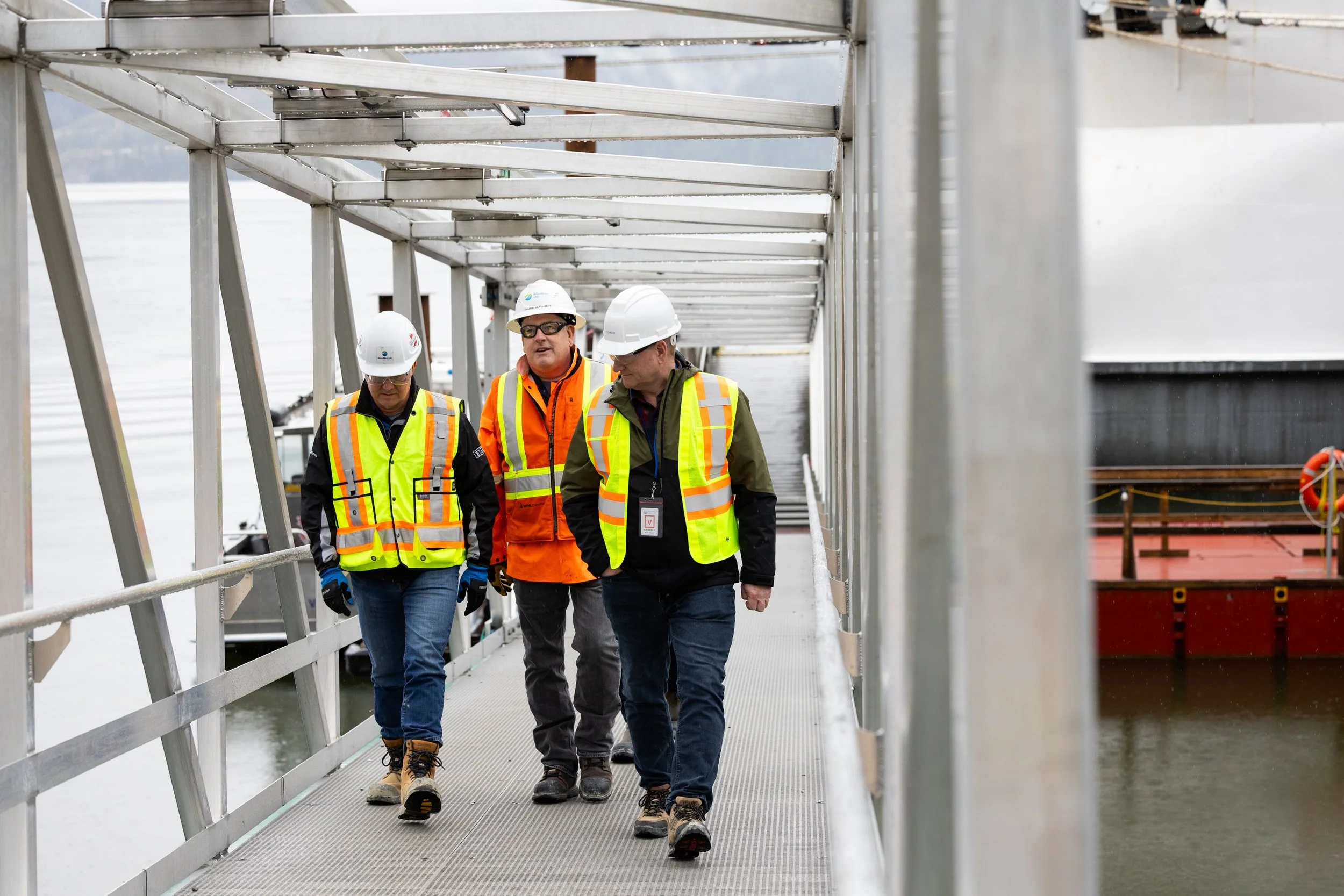Three workers wearing safety helmets and reflective vests walk on a metal platform over water, possibly at a dock or construction site.