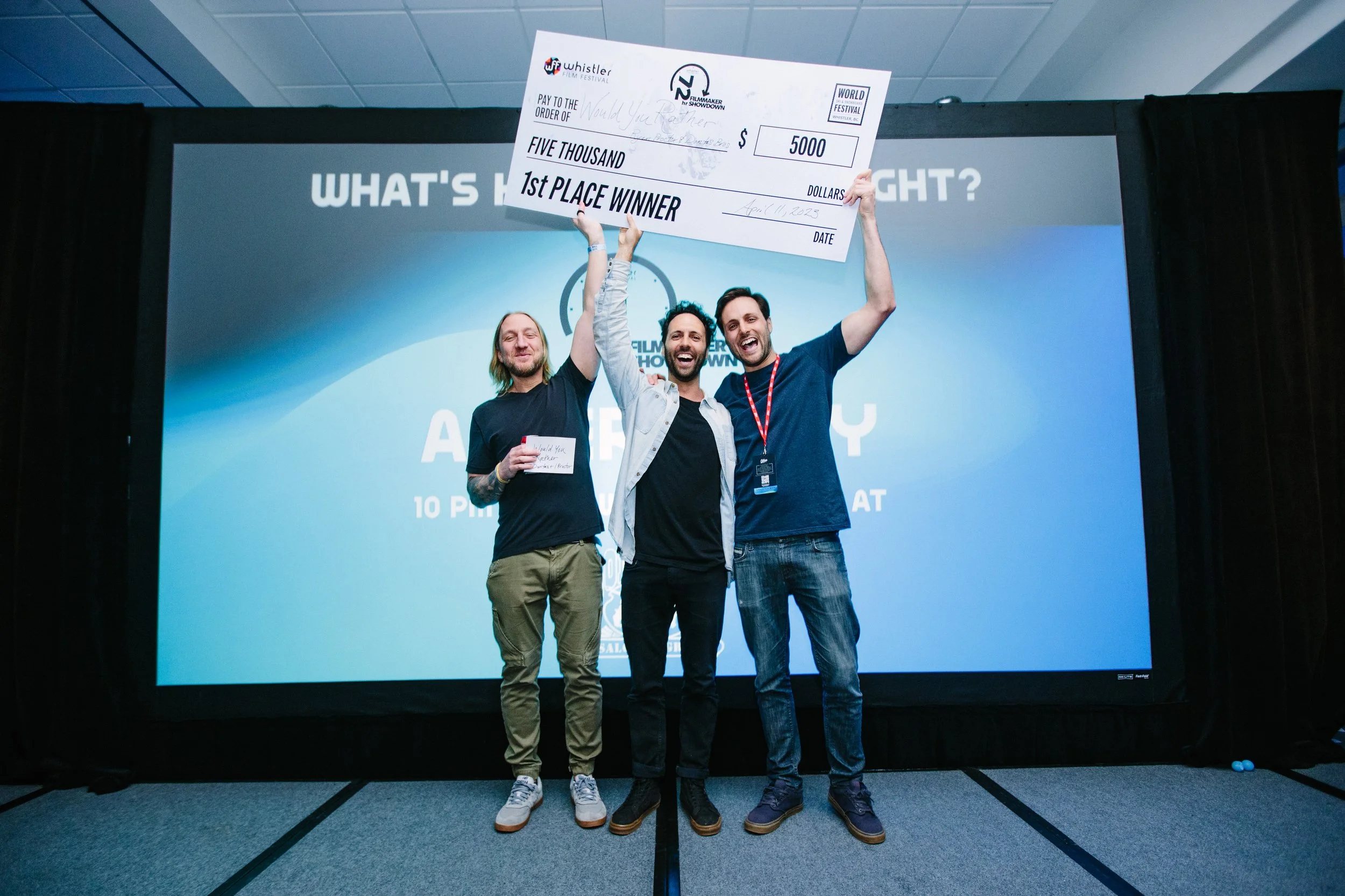 Three men on stage celebrating, holding a large check for $5000, with a screen in the background displaying 'WHAT'S YOUR STORY?'}