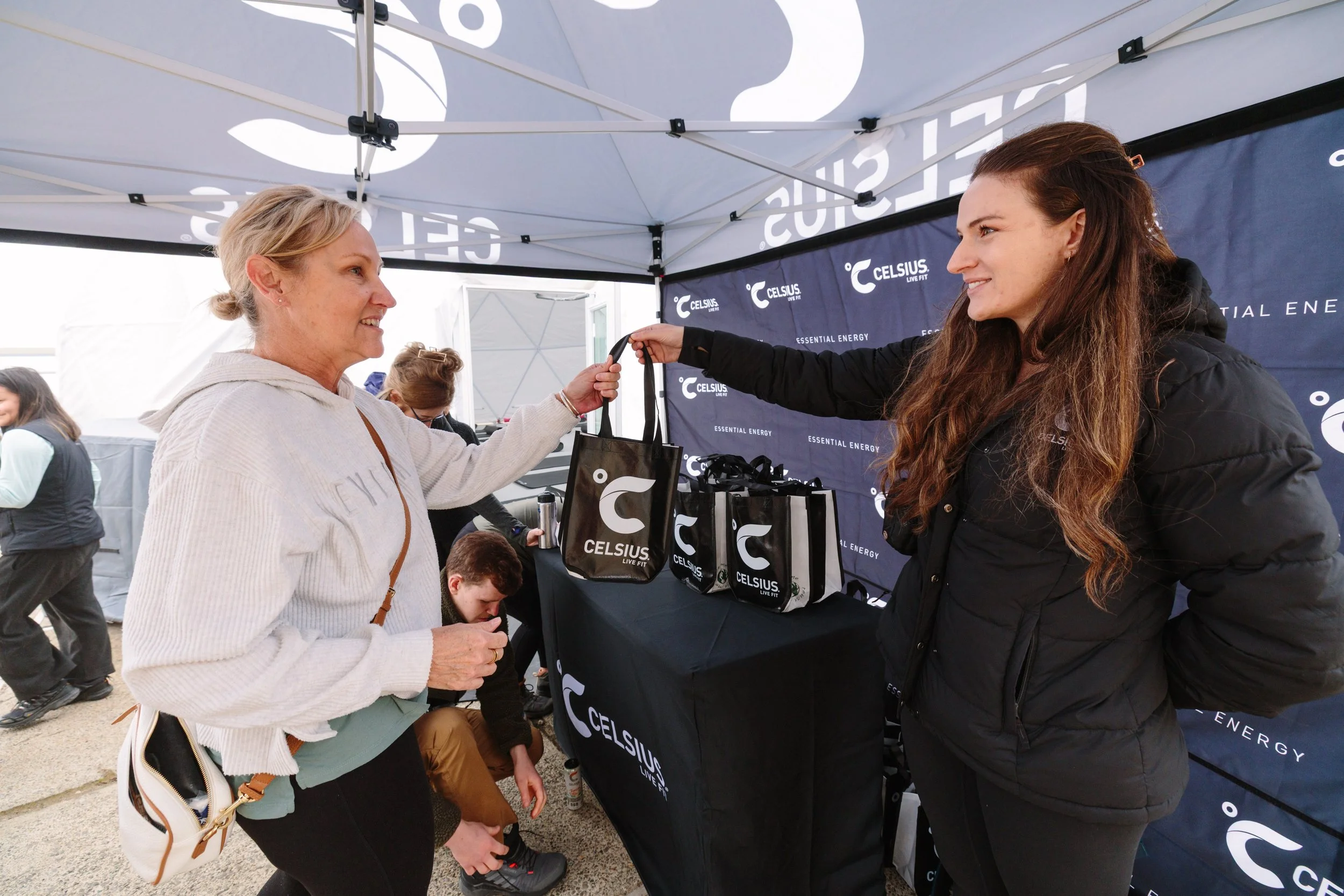 A woman receiving a black bag from a sales representative at a Celsius Energy booth during an outdoor event.