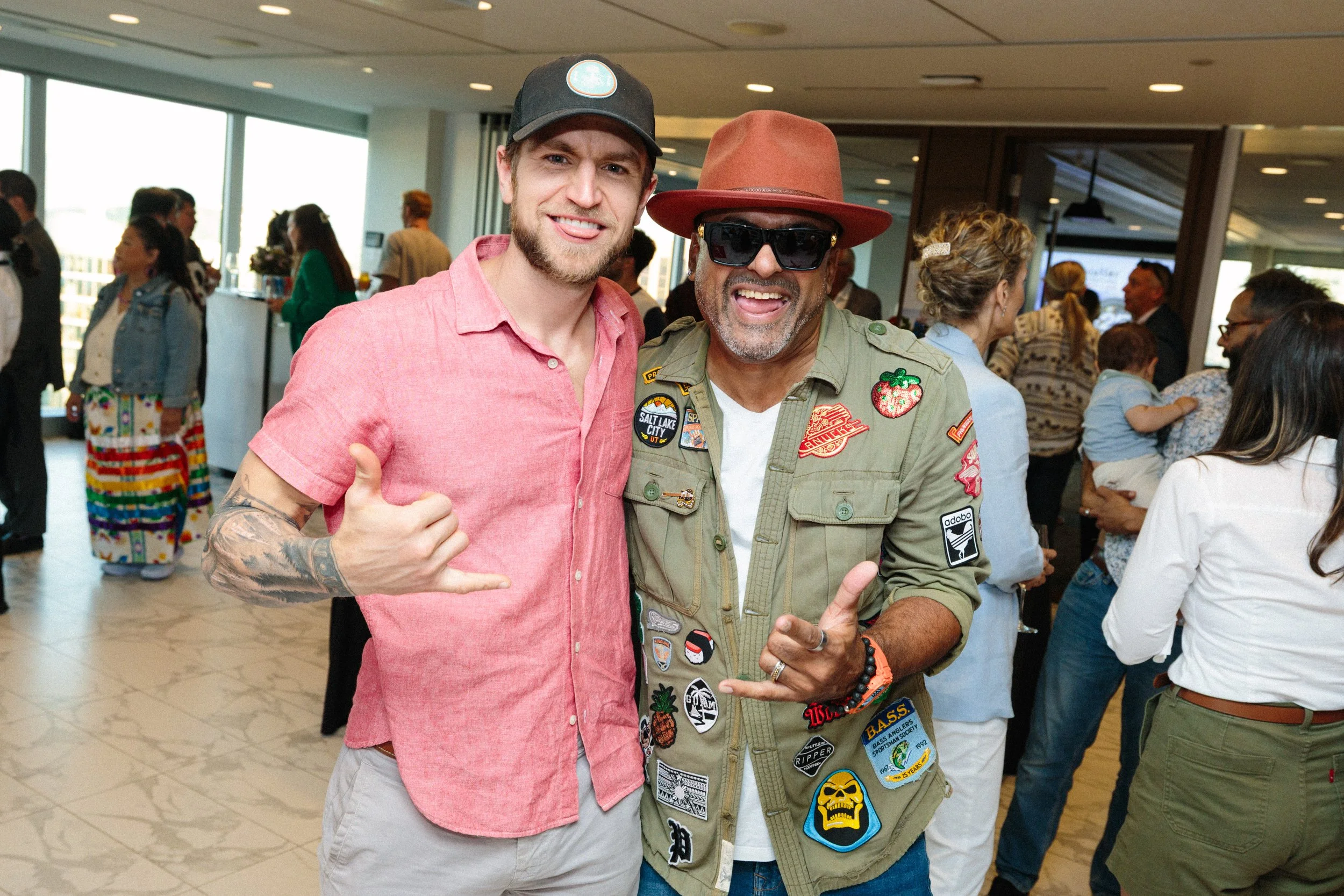 Two men smiling and making shaka signs at an indoor social gathering, with people in the background engaging and chatting.
