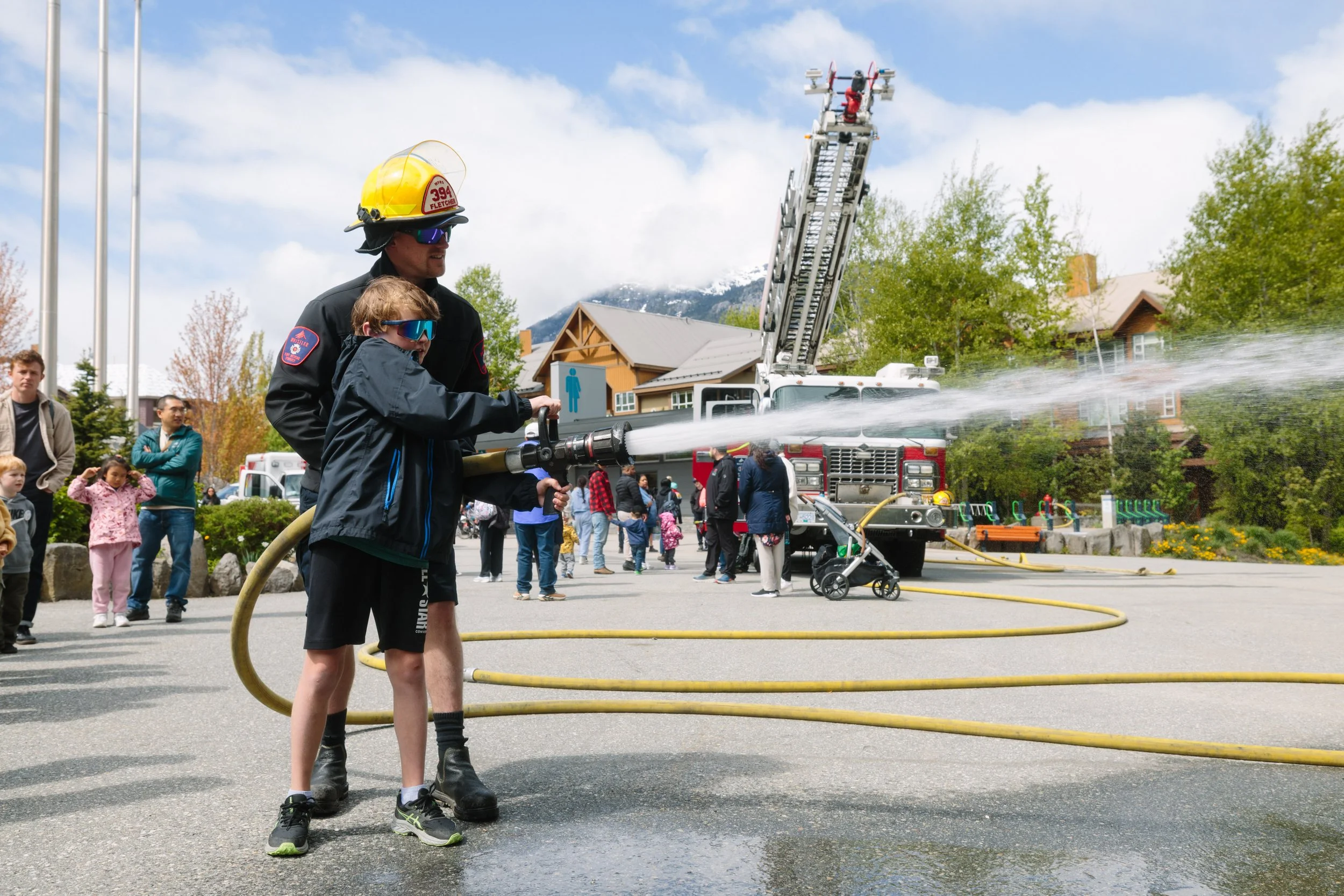 A firefighter teaches a young boy how to hold and spray a fire hose at a public safety event, with onlookers and fire trucks in the background.