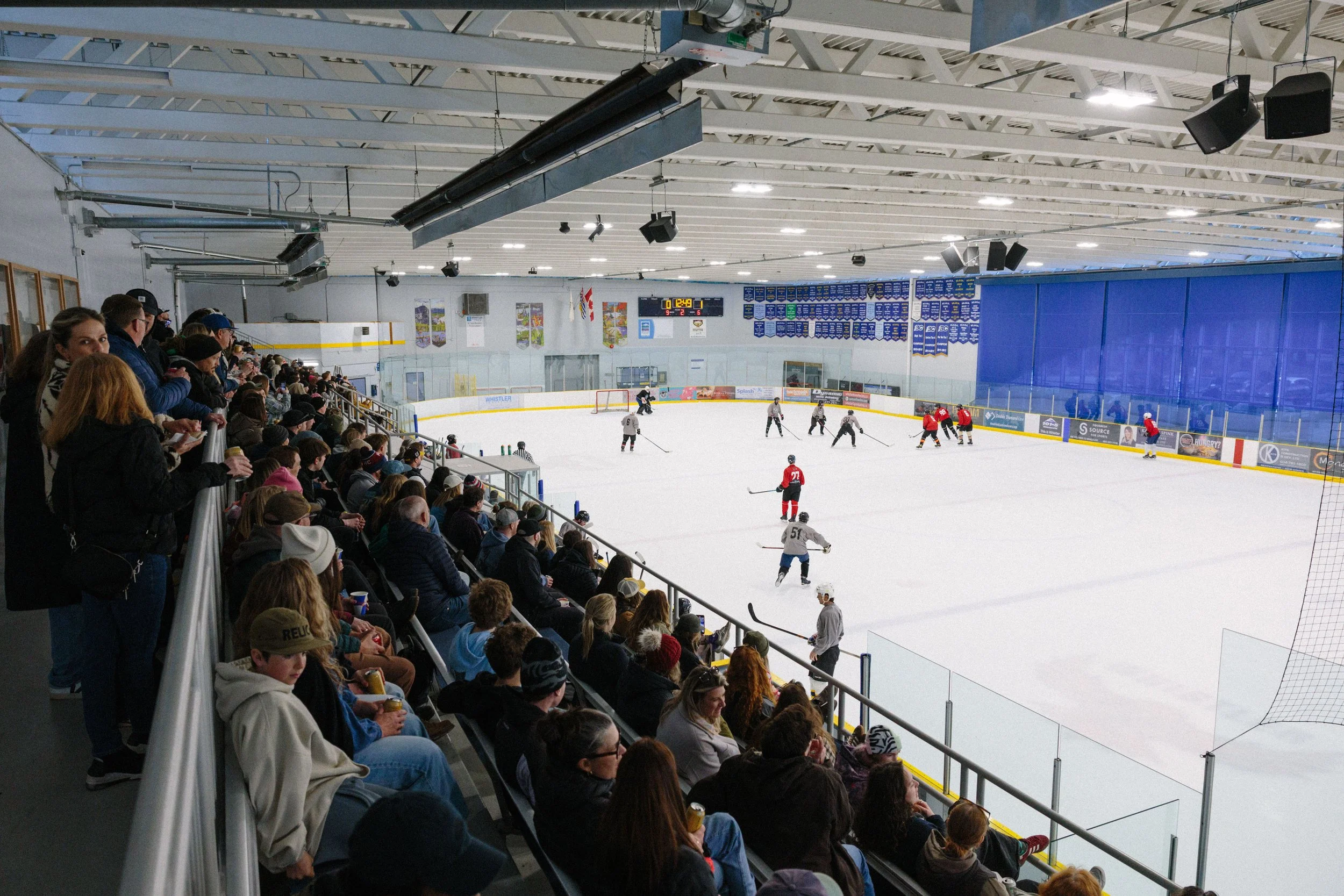 An indoor ice hockey game with spectators in the stands. Players are on the ice, some wearing white and others in red uniforms. The arena has banners on the walls and a digital scoreboard displaying the game time and scores.