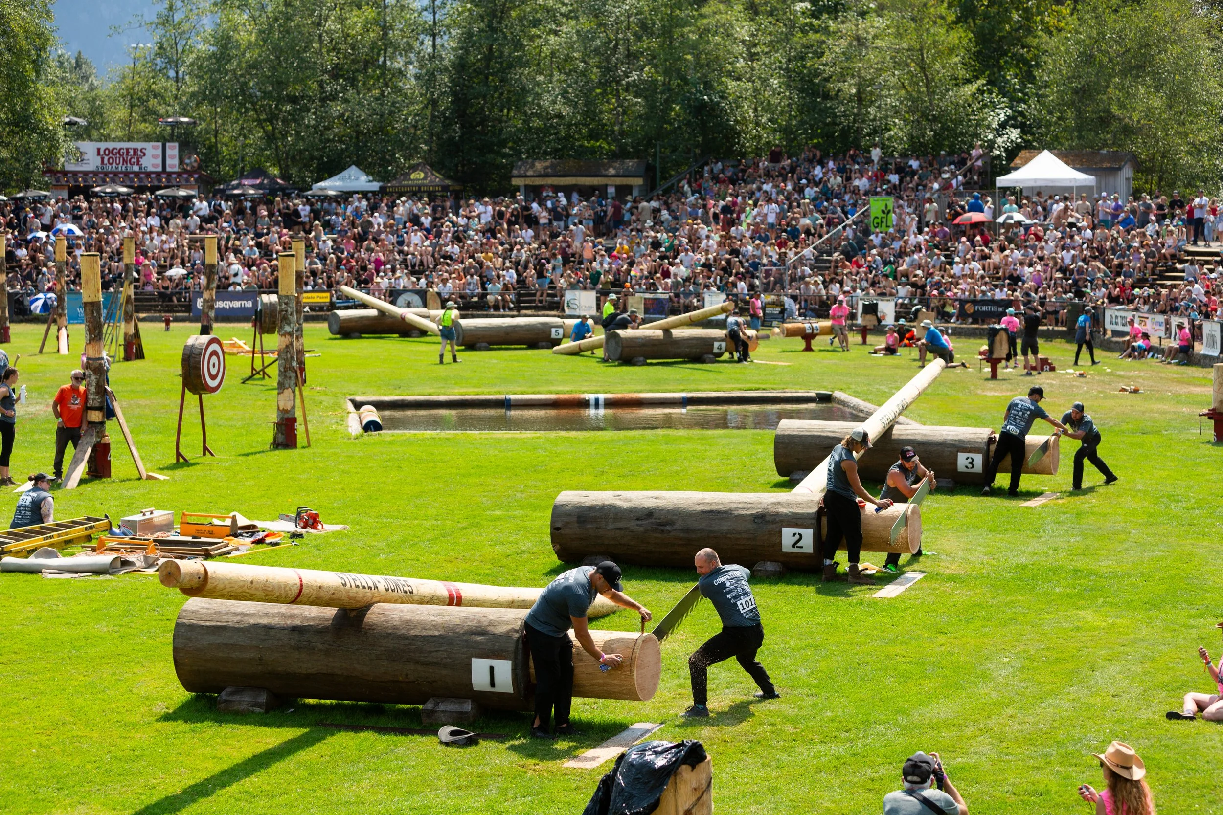 A large outdoor event focused on lumberjack competitions, with contestants chopping logs and performing sawmill tasks on a grassy field, filled with spectators in bleachers and around the area.