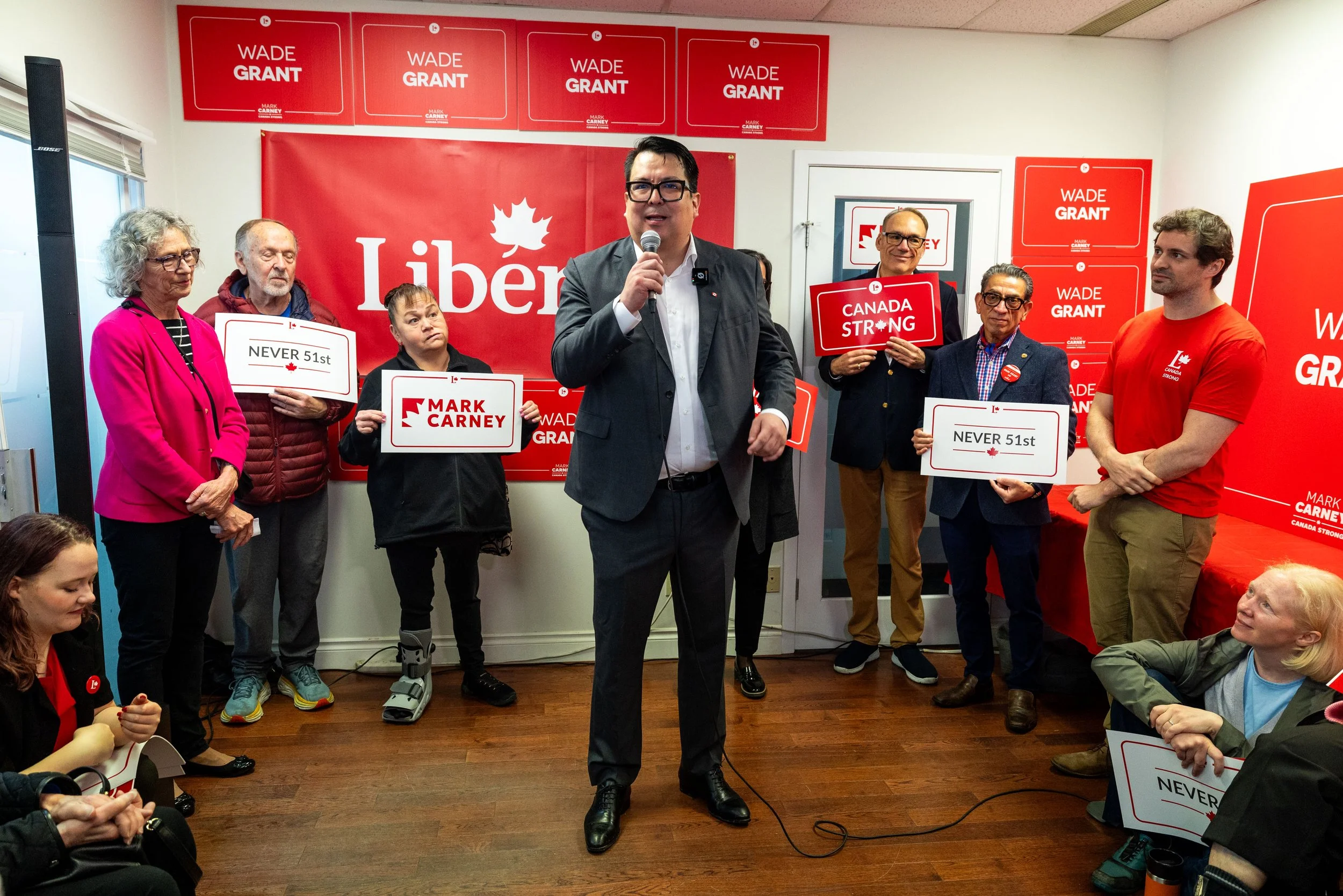 A group of people standing indoors at a political event, with a man in a gray suit holding a microphone in the foreground, and others holding signs for Wade Grant, Mark Carney, and Canada Strong, against a red backdrop with political branding.