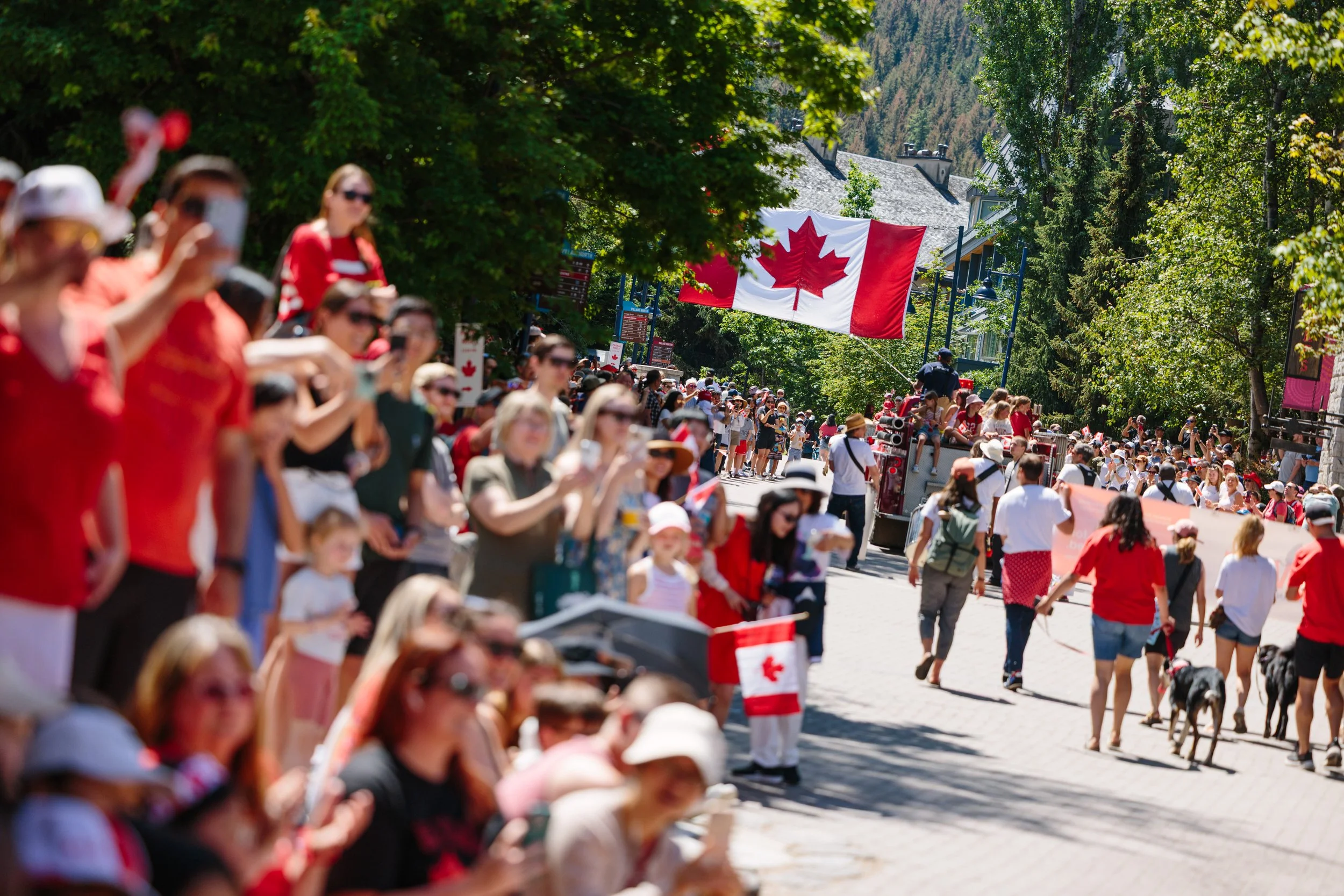 Crowd of people gathering outdoors for a celebration with Canadian flags and red clothing, surrounded by green trees and a Canadian flag on a float.