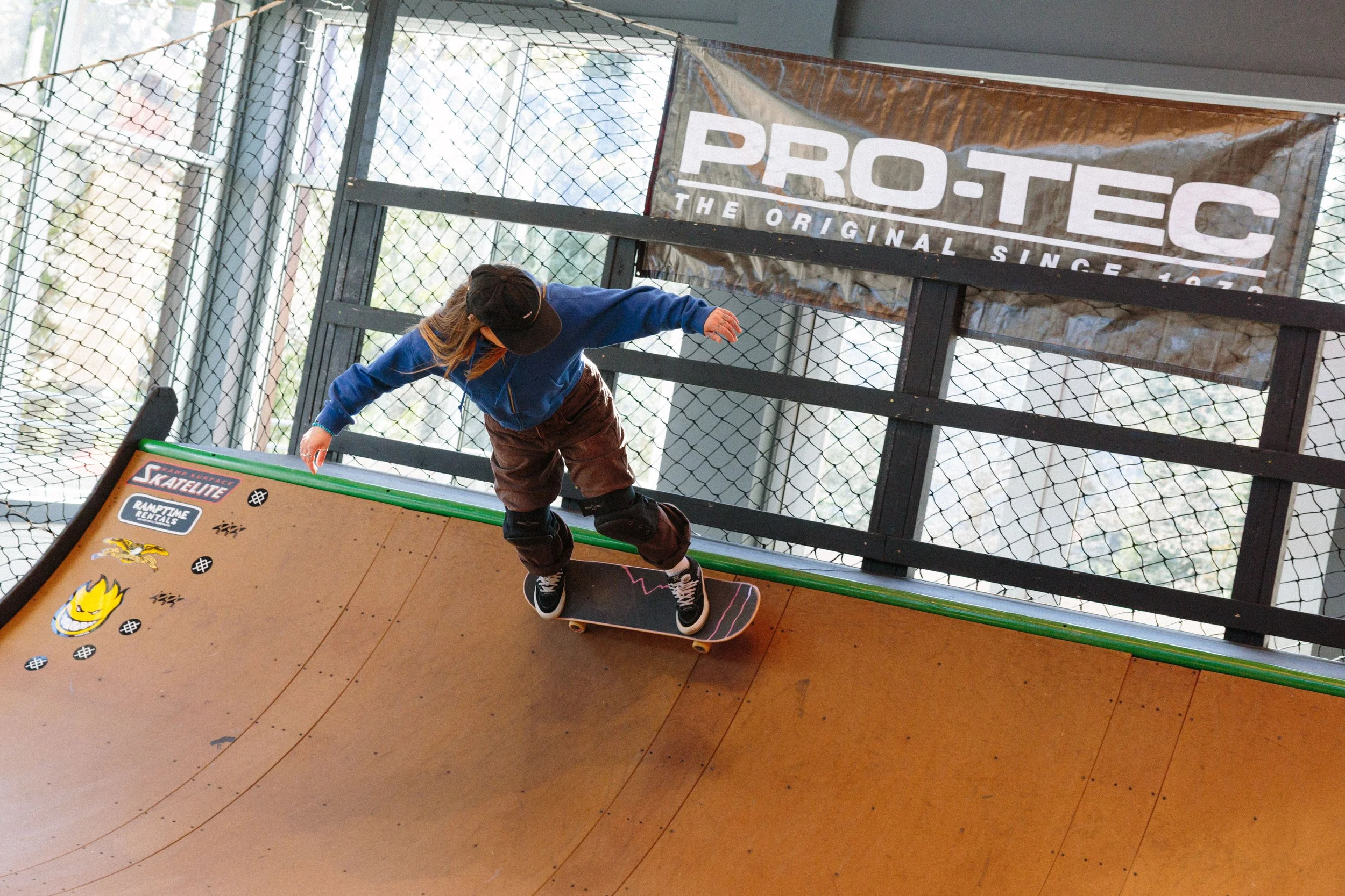 A young person skateboarding indoors, wearing a black cap, blue sweatshirt, brown pants, and black sneakers, on a skateboard with pink accents. There is a large sign that reads 'PRO-TEC THE ORIGINAL SINCE,' and the background includes a chain-link fe