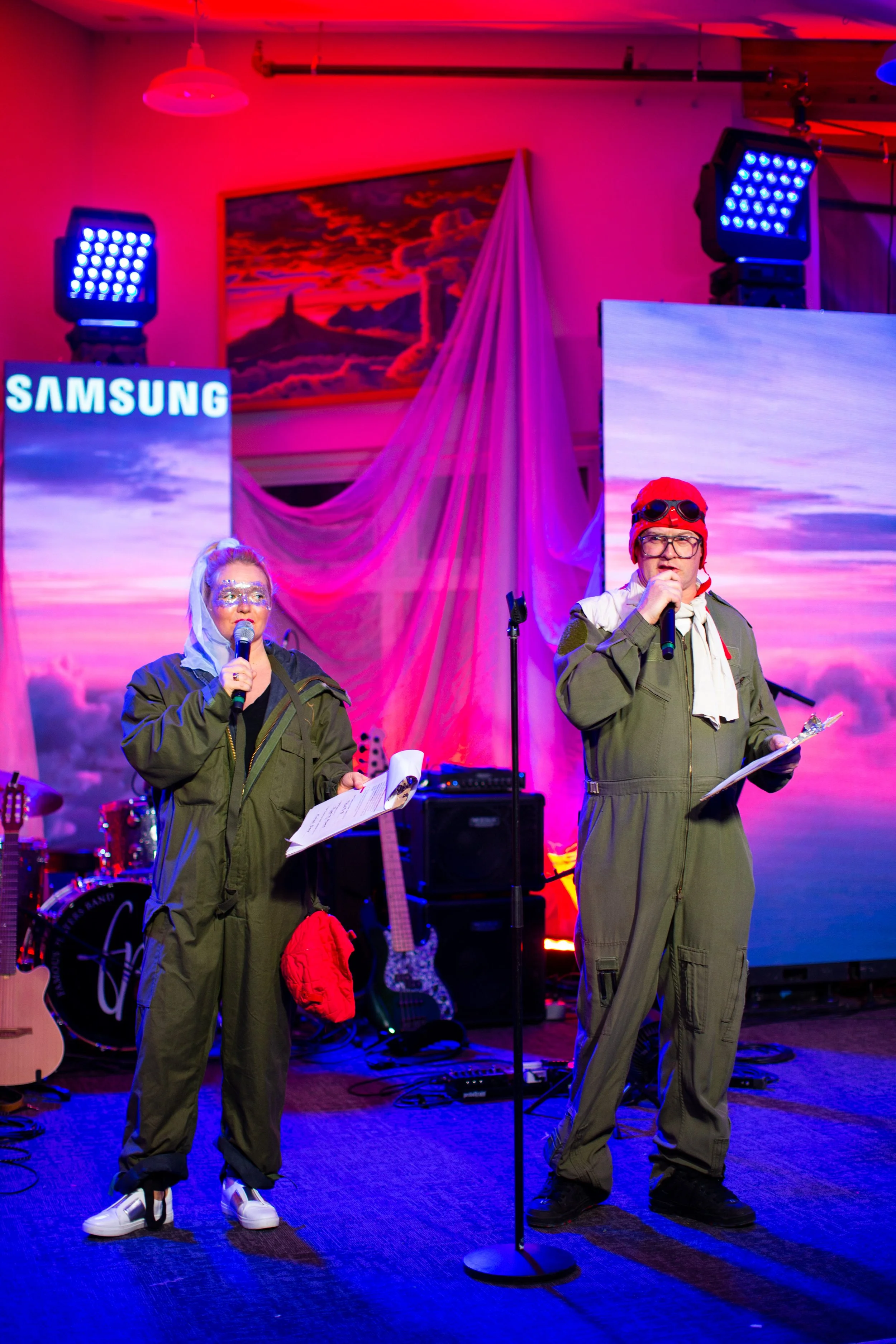 Two performers dressed in military-style outfits singing into microphones on a brightly lit stage with musical instruments and vibrant pink and purple background visuals.