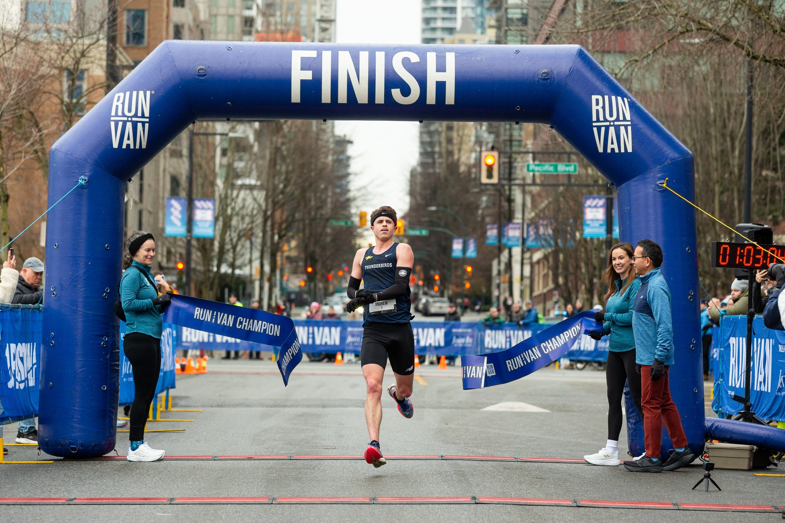 A male runner crossing the finish line at a race, with an inflatable blue arch marked 'FINISH' and surrounded by spectators and race officials.