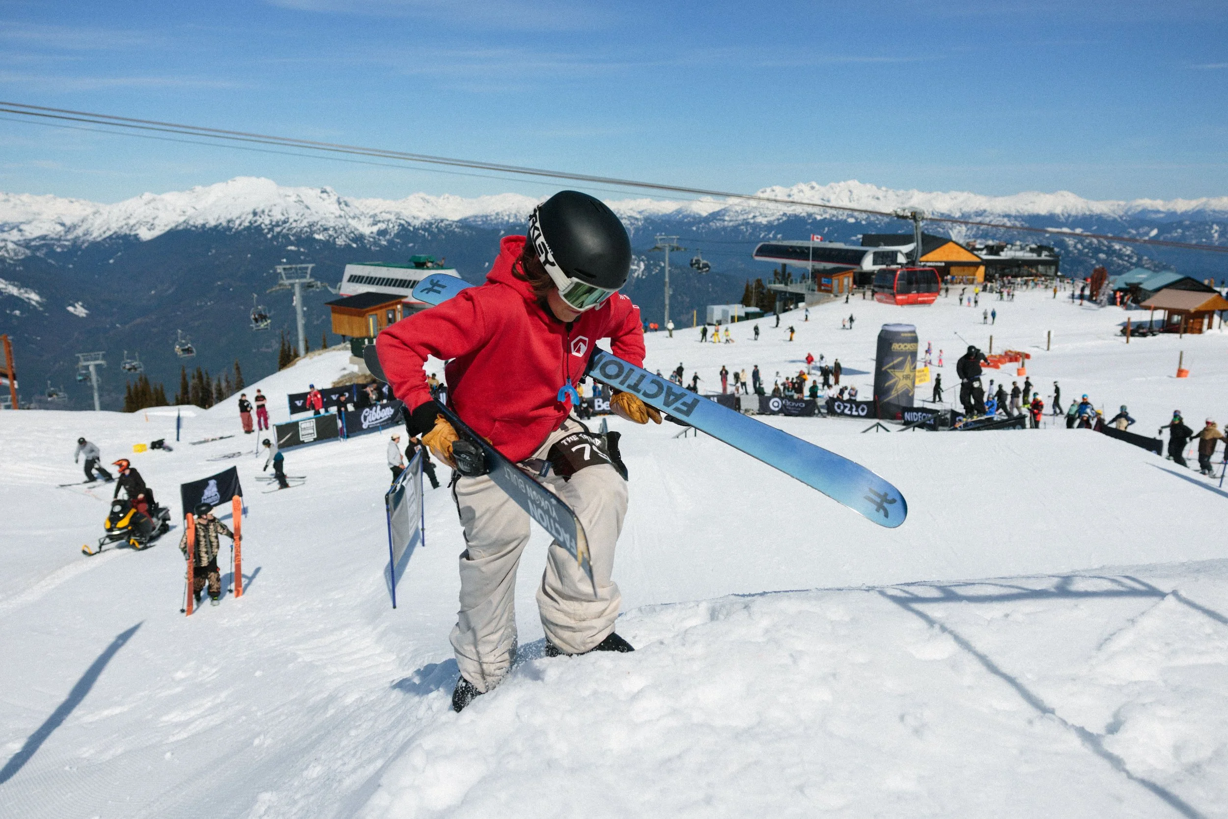A skier in a red jacket and beige pants adjusting skis on a snowy slope at a ski resort, with ski lifts and other skiers in the background, mountains with snow-capped peaks, and a clear blue sky.
