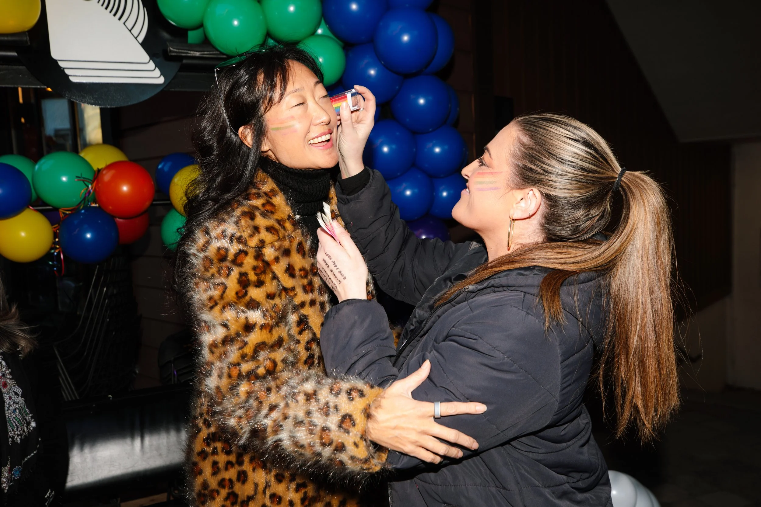 Two women celebrating at a party, one is applying face paint to the other, with colorful balloons in the background.