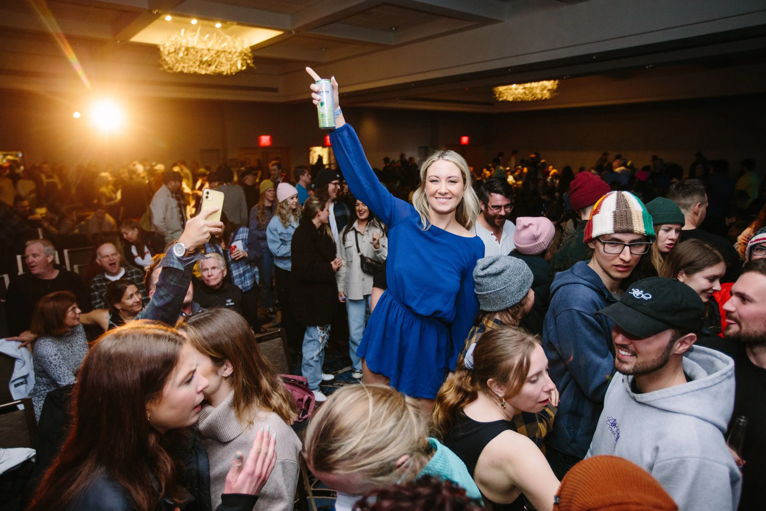A smiling woman in a blue dress standing amidst a crowded indoor event, raising a can of Corona beer in her right hand.