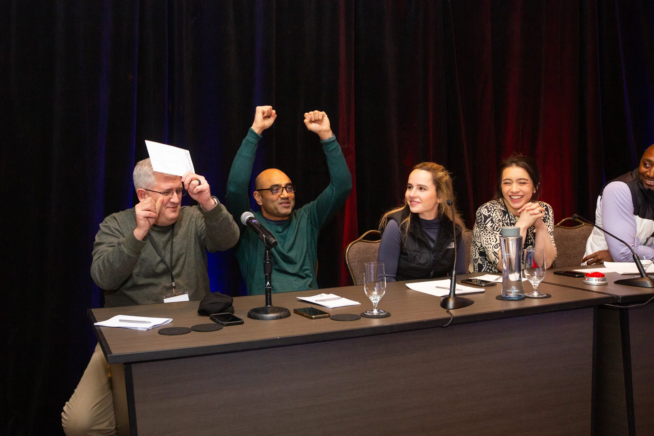Group of five people sitting at a panel table, one holding a paper, others smiling and gesturing with joy, microphones in front.