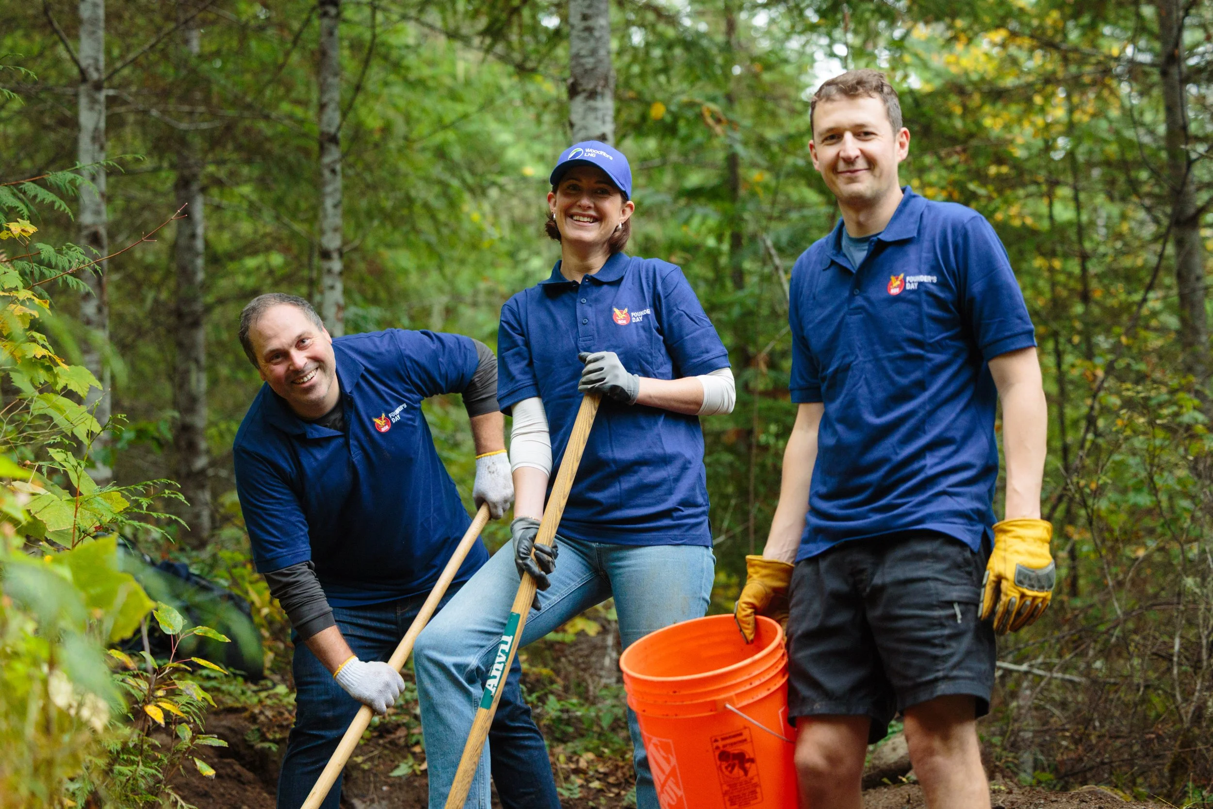 Group of three volunteers in blue shirts working outdoors in a wooded area during daytime, holding shovels and an orange bucket, smiling and posing for the photo.