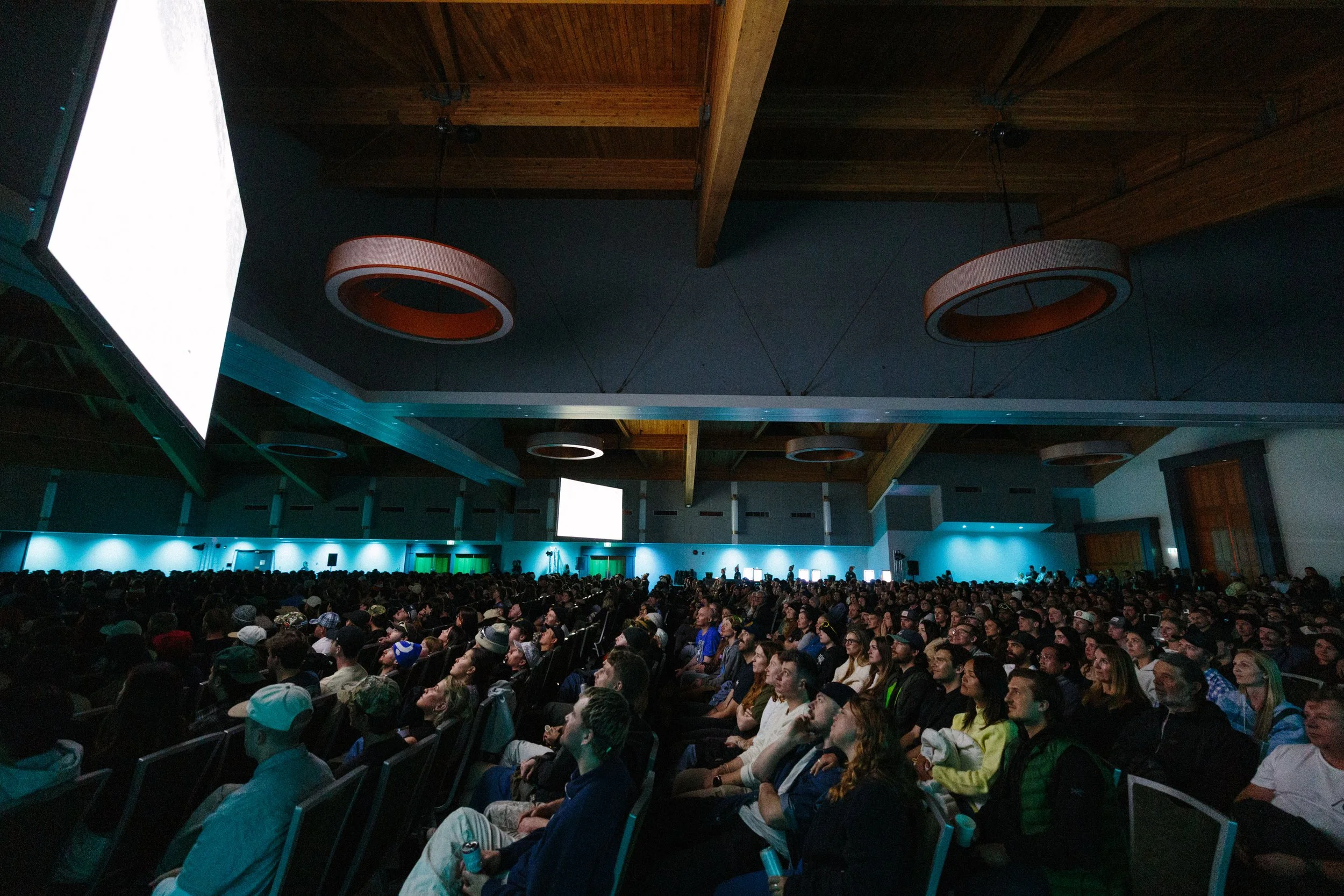 A large indoor event with a crowd of people seated facing a stage, with a dark ceiling featuring hanging circular light fixtures and wooden beams, and a bright rectangular light panel on the left side.