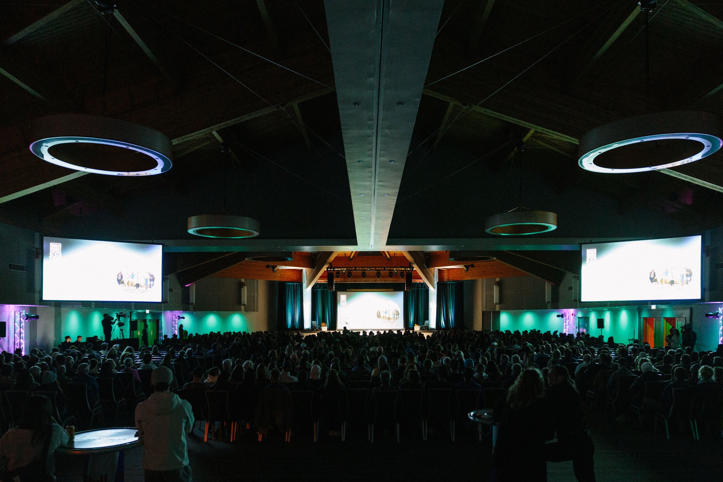A large audience seated in a dark auditorium watching a presentation with a large screen on stage. The stage has multiple screens displaying visuals, and there are circular lights hanging from the ceiling.