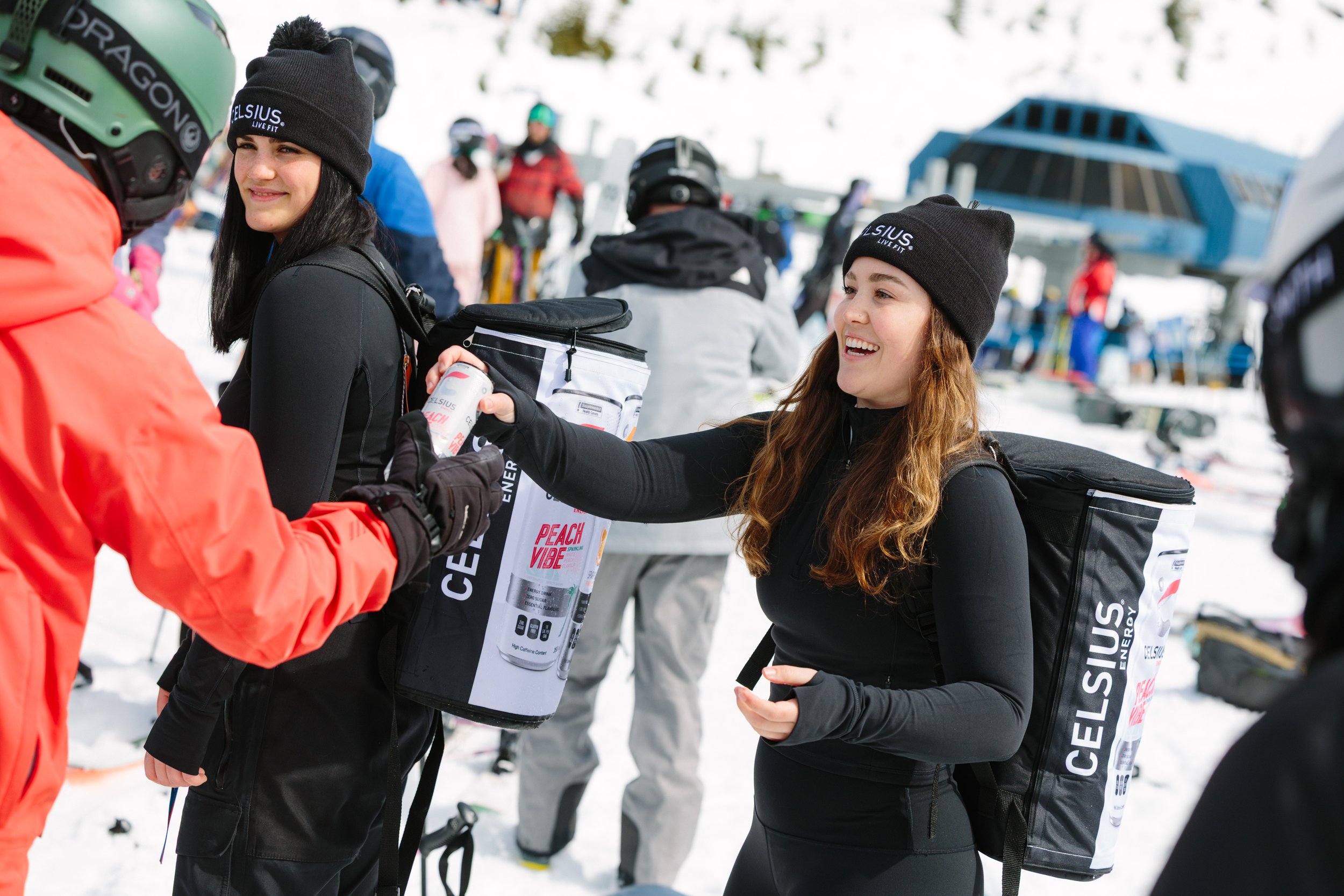 Two women in black winter clothing and hats exchanging bottles at a snowy ski resort, with skiers and a ski lift in the background.
