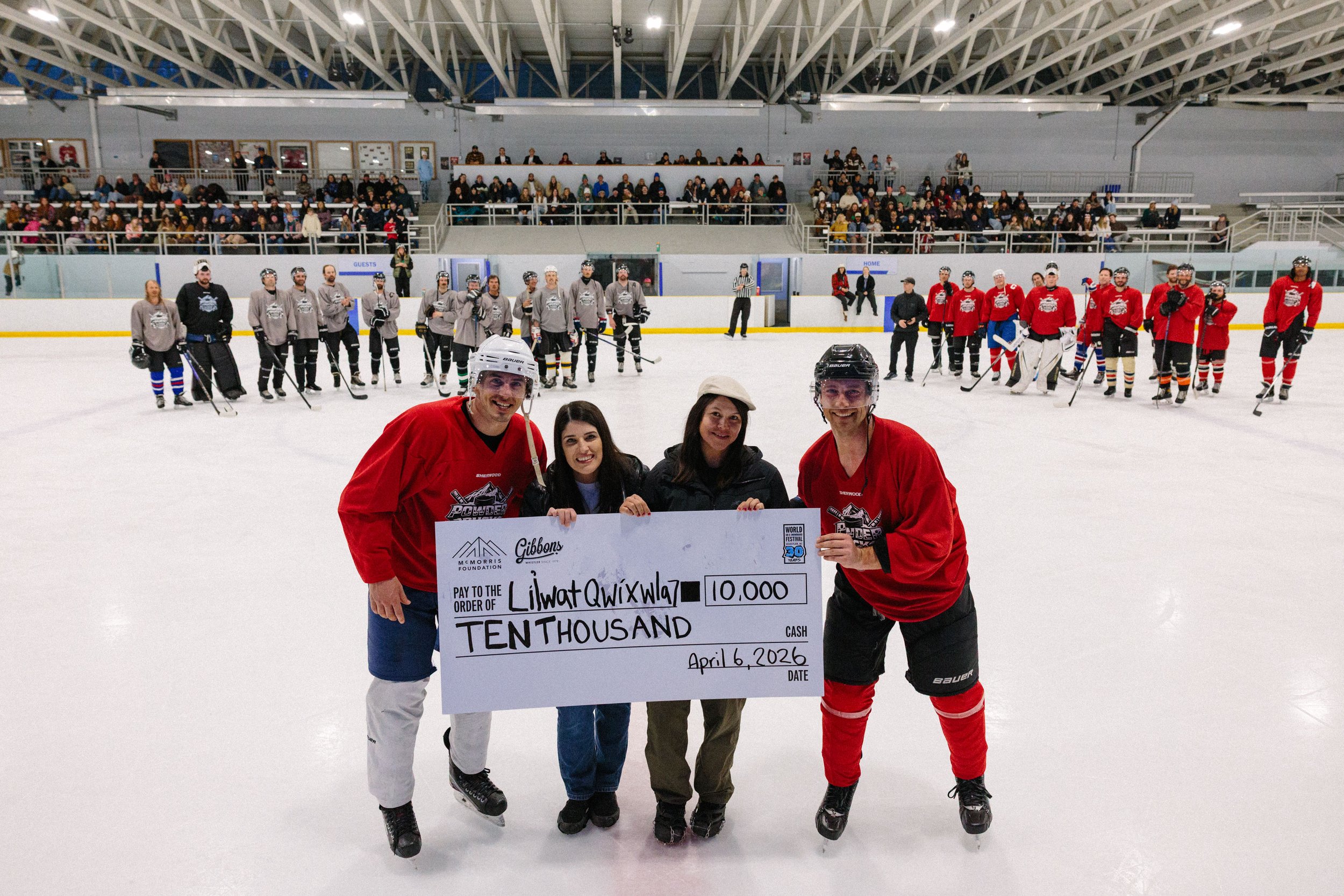 Four people are holding a large check on an indoor ice rink, with hockey players and spectators in the background.