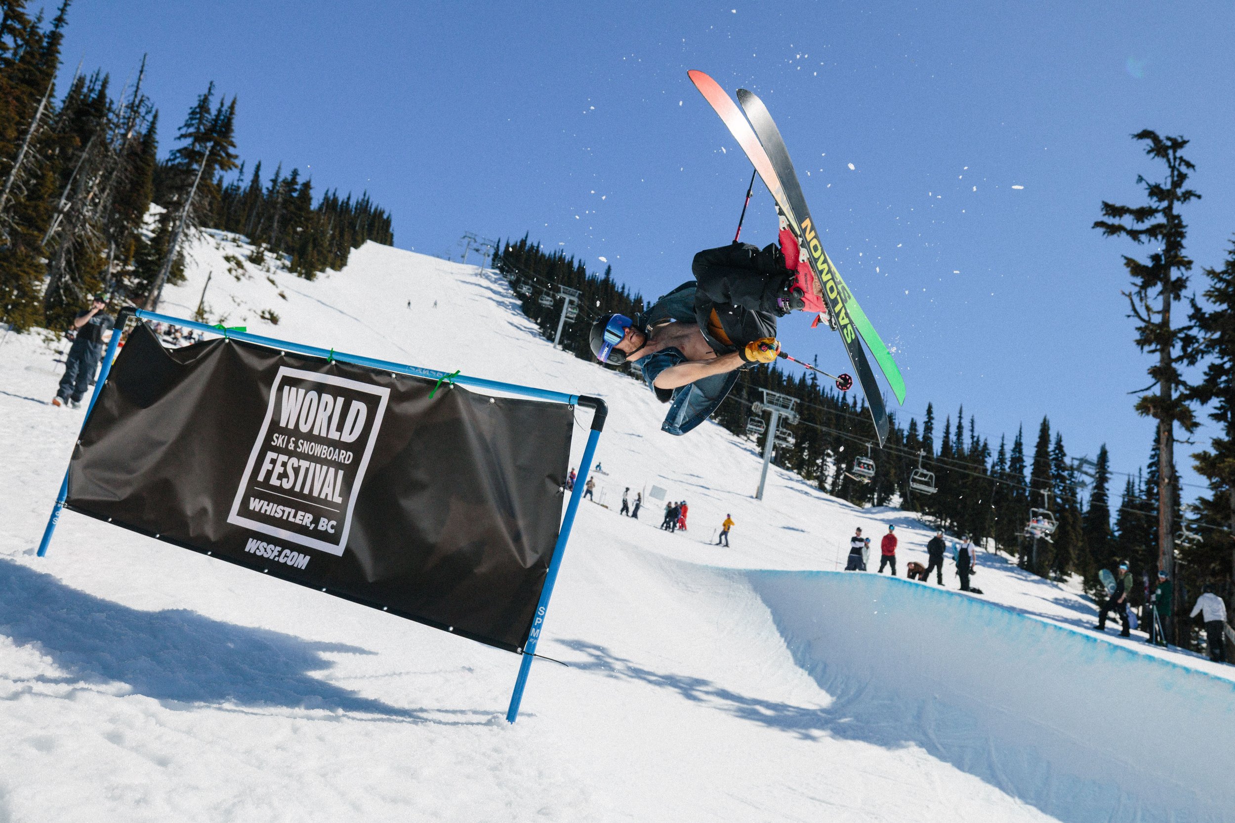 A skier performing a jump over a banner at a ski and snowboard festival in Whistler, BC, on a sunny day with a snowy mountain and ski lift in the background.