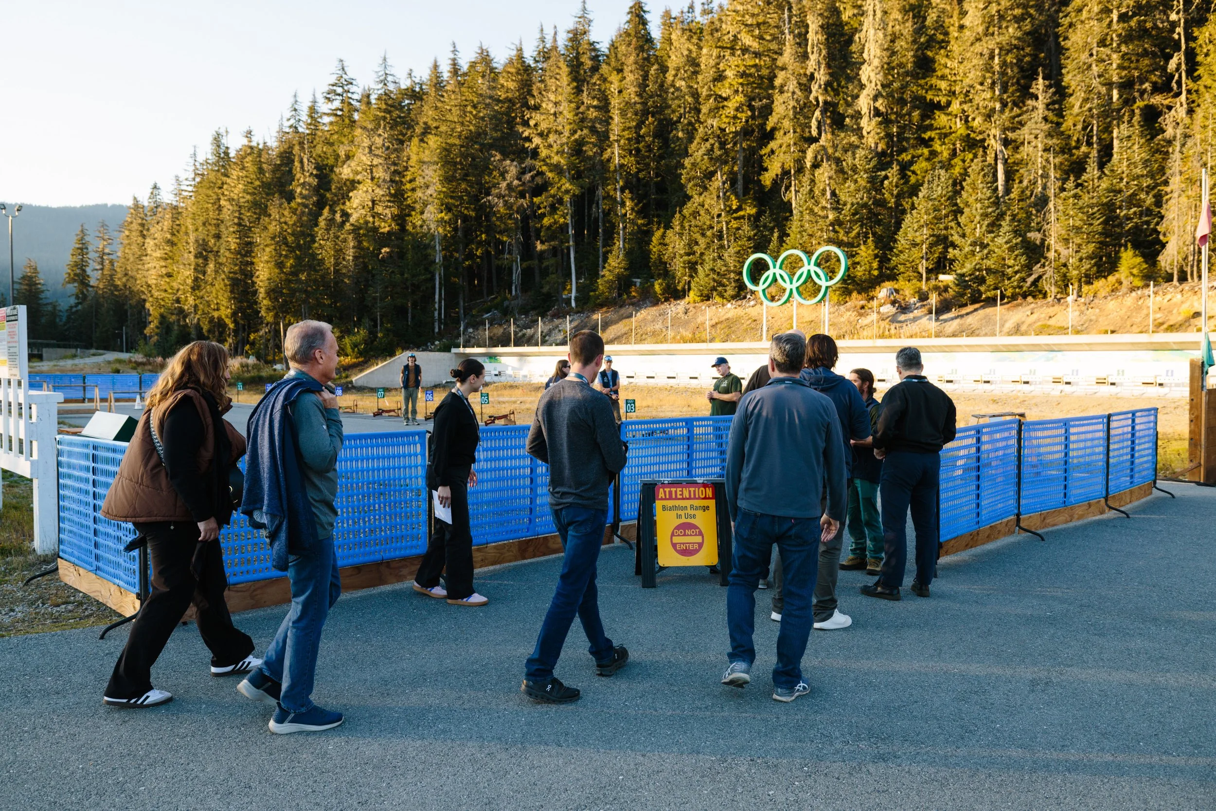 Line of people waiting outside a biathlon range with blue barriers, a yellow sign, and a backdrop of tall pine trees and a mountain range, in the late afternoon.