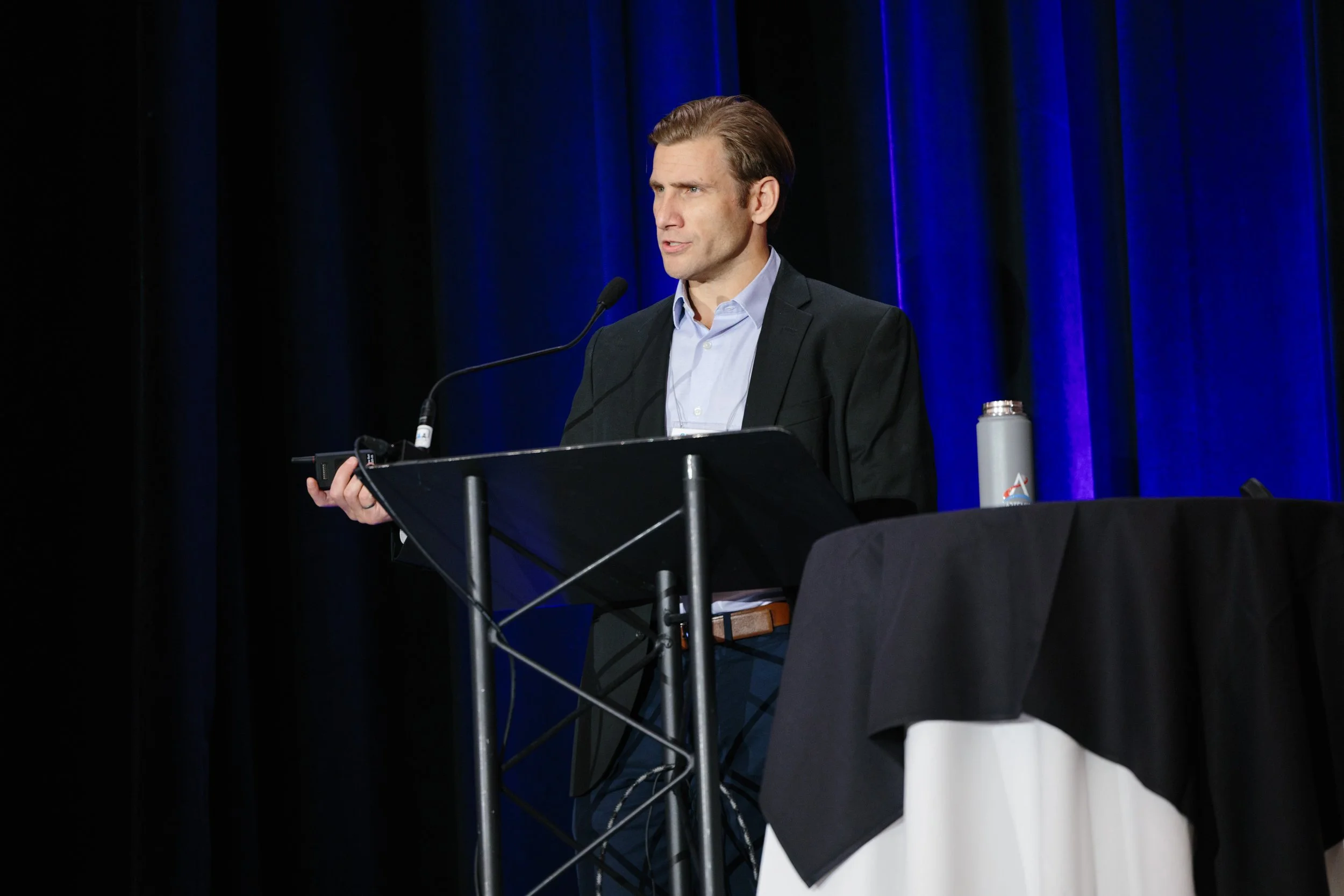 A man in a suit speaking at a podium during a conference or presentation, with a blue curtain background and a table with a water bottle on it.