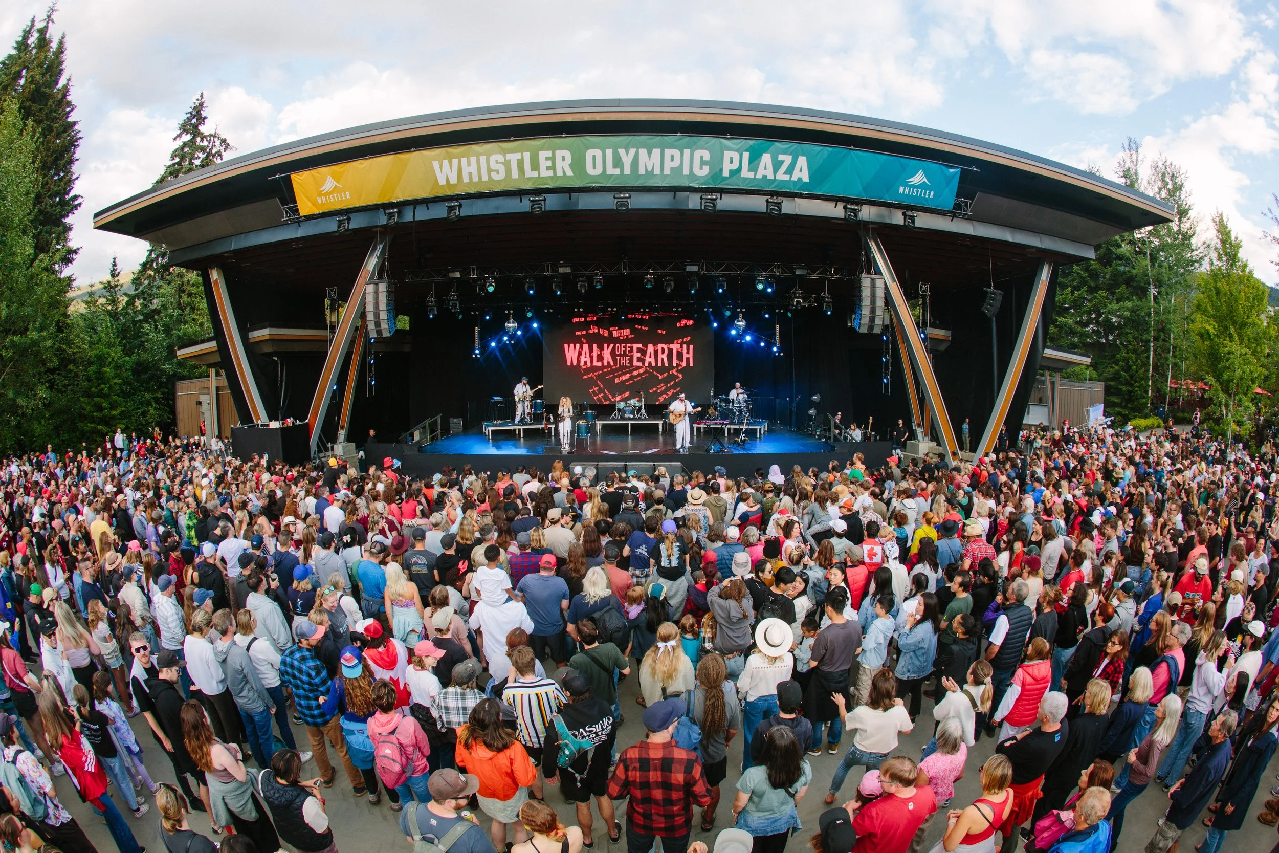 Large crowd attending an outdoor concert at Whistler Olympic Plaza with a stage performance for the event 'Walk of the Earth'.