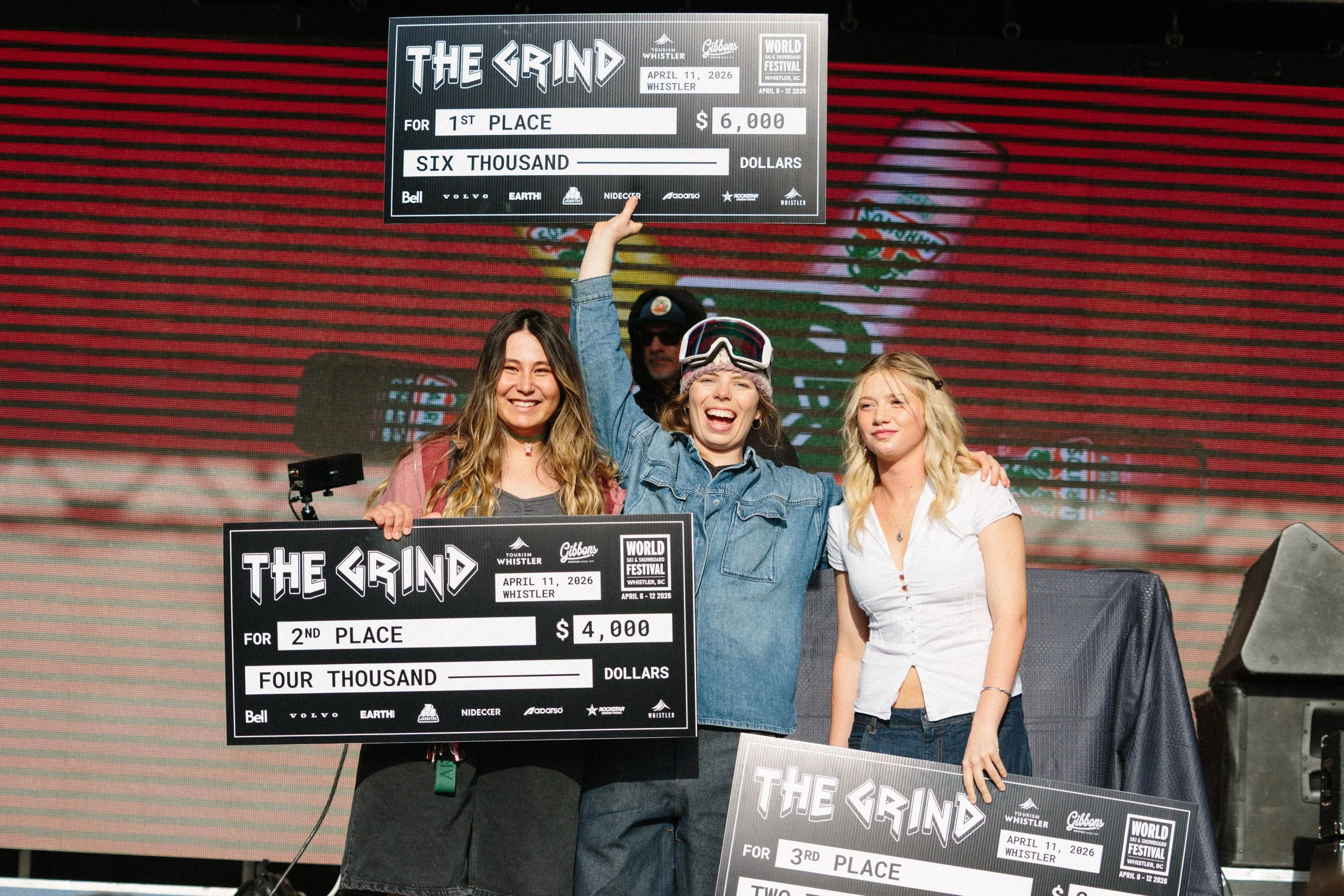 Three women on stage holding large checks for a snowboarding event in Whistler, Canada. The woman in the middle is smiling, wearing ski goggles on her head, and raising her right arm. The women on either side are smiling, with the woman on the right 
