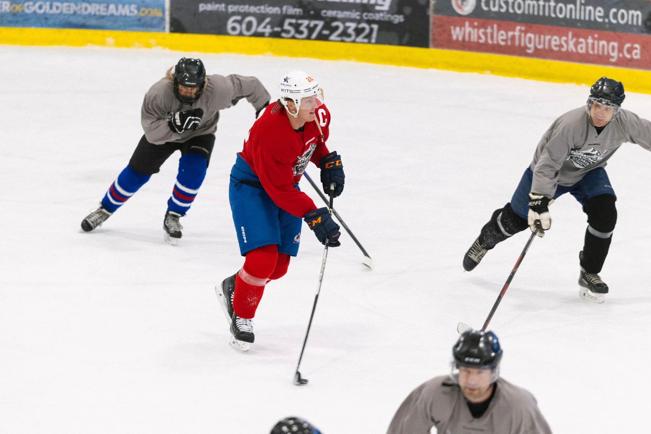 Hockey players on ice rink, one wearing red jersey with Captain C, others in gray jerseys, wearing helmets and skates, holding hockey sticks, with banners and advertisements on the wall in the background.