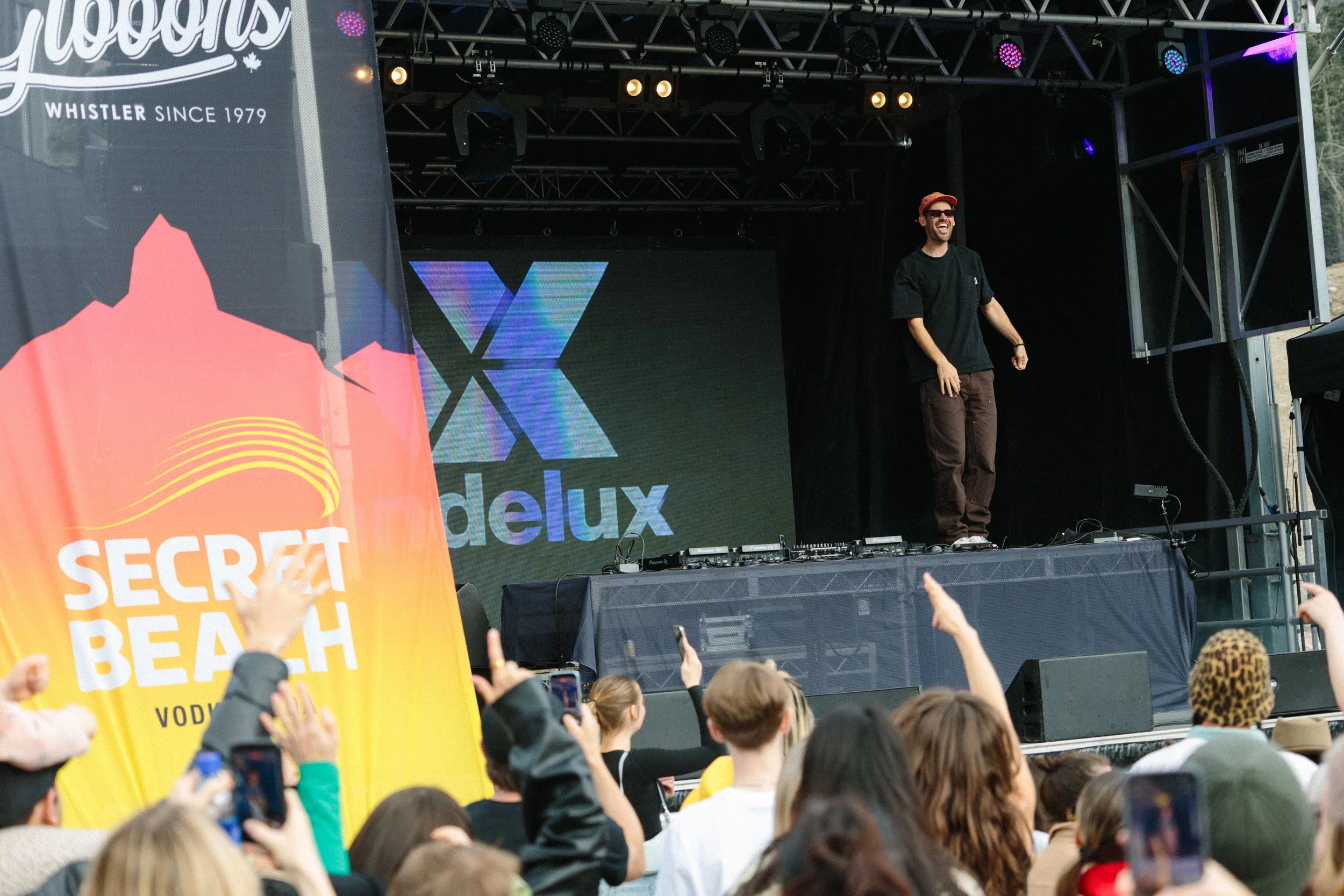 A man wearing sunglasses and a hat stands on an outdoor stage at a music event, with colorful lights and a screen behind him displaying the word 'deluxe.' A crowd is visible in front of the stage, with some holding up their phones and a bright yellow