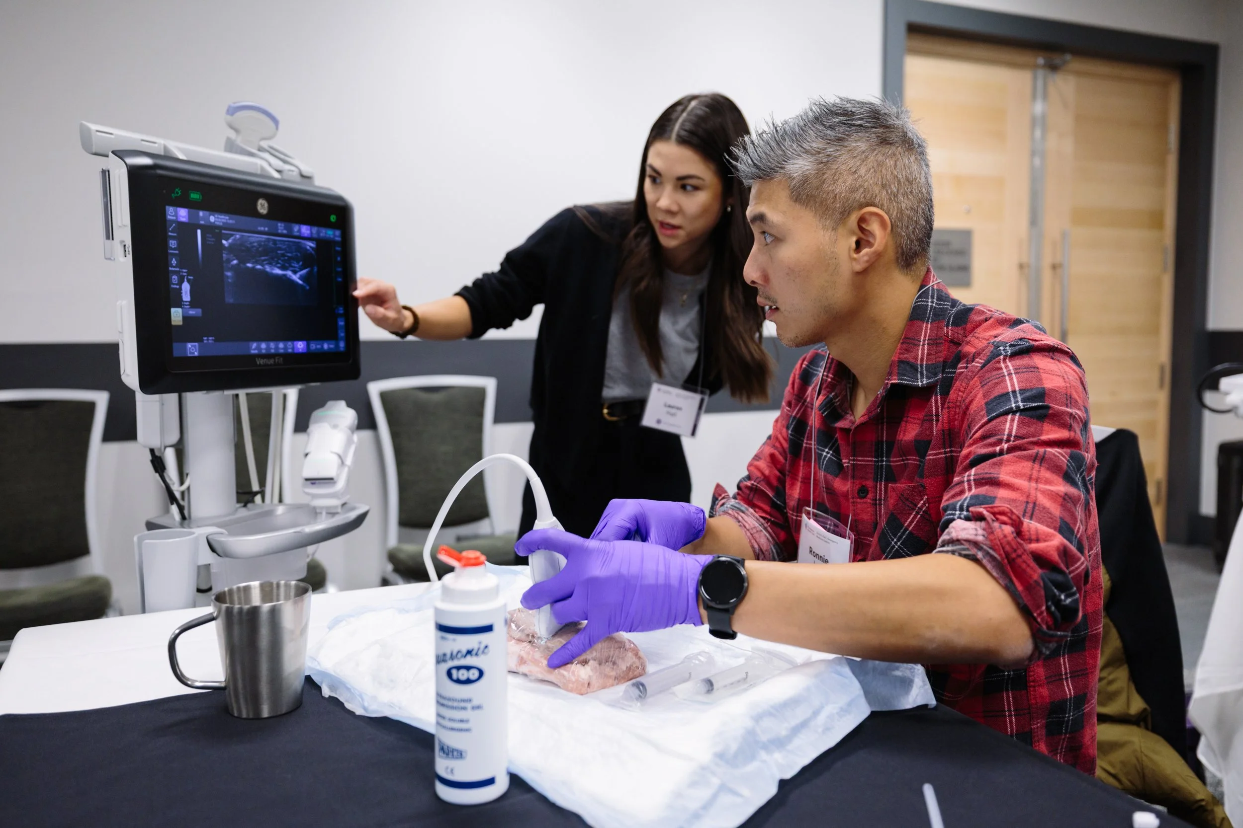 A man wearing purple gloves and a red plaid shirt practices a medical procedure using a model of a human lung and an ultrasound machine, while a woman in black observes and explains, in a training or workshop setting.