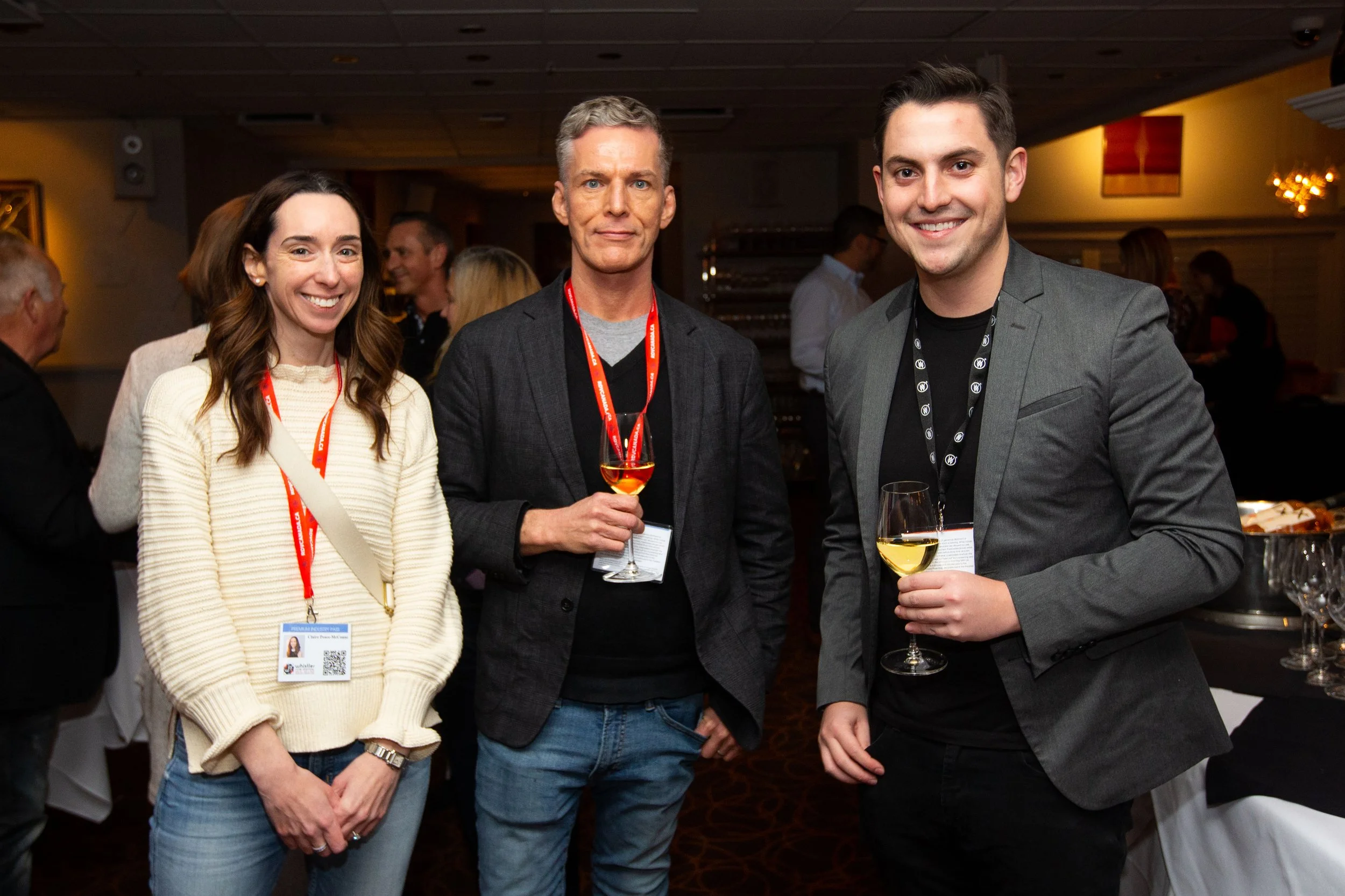 Group of three people at a social event holding wine glasses, wearing lanyards.