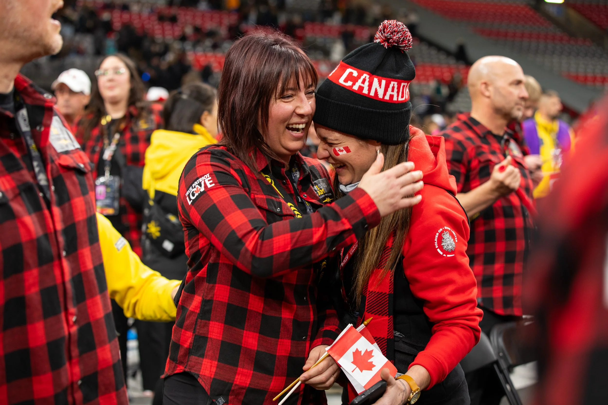 Two women wearing red and black plaid shirts, one with a Canada hat and face paint, embracing and smiling at a sports event, holding a small Canadian flag.