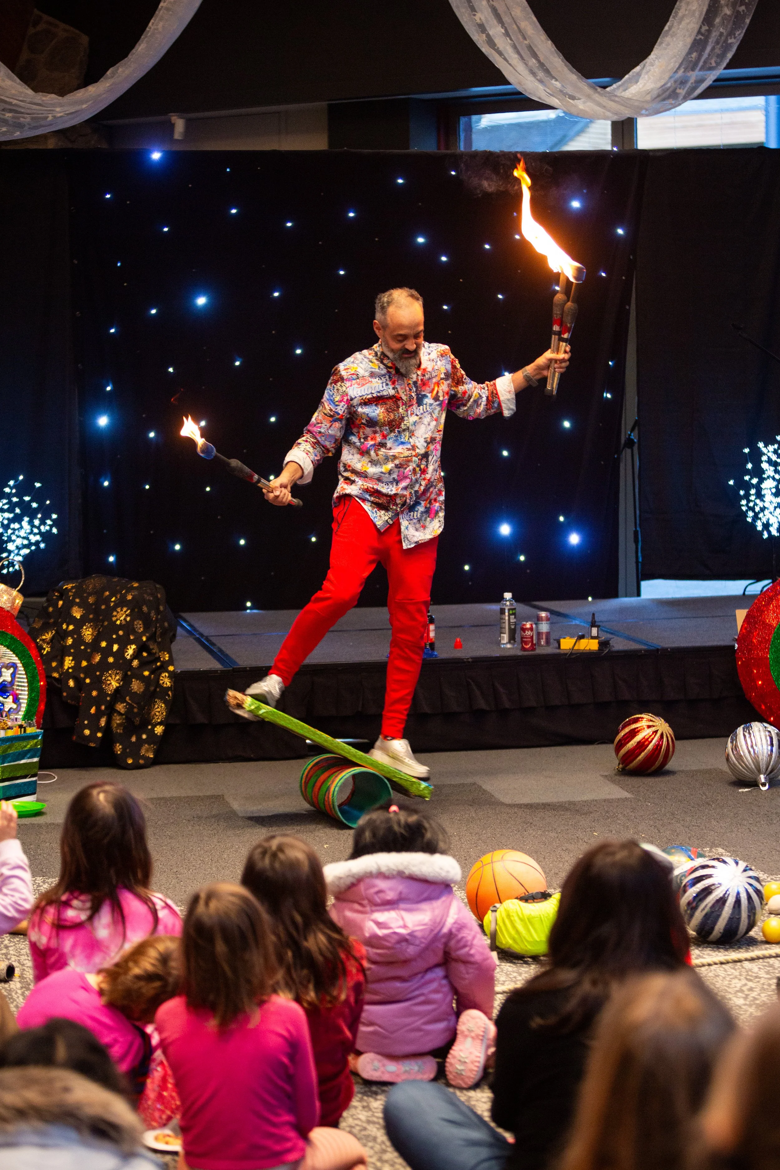 Magician performing with fire juggling on stage in front of children, decorated with Christmas ornaments and festive decorations.