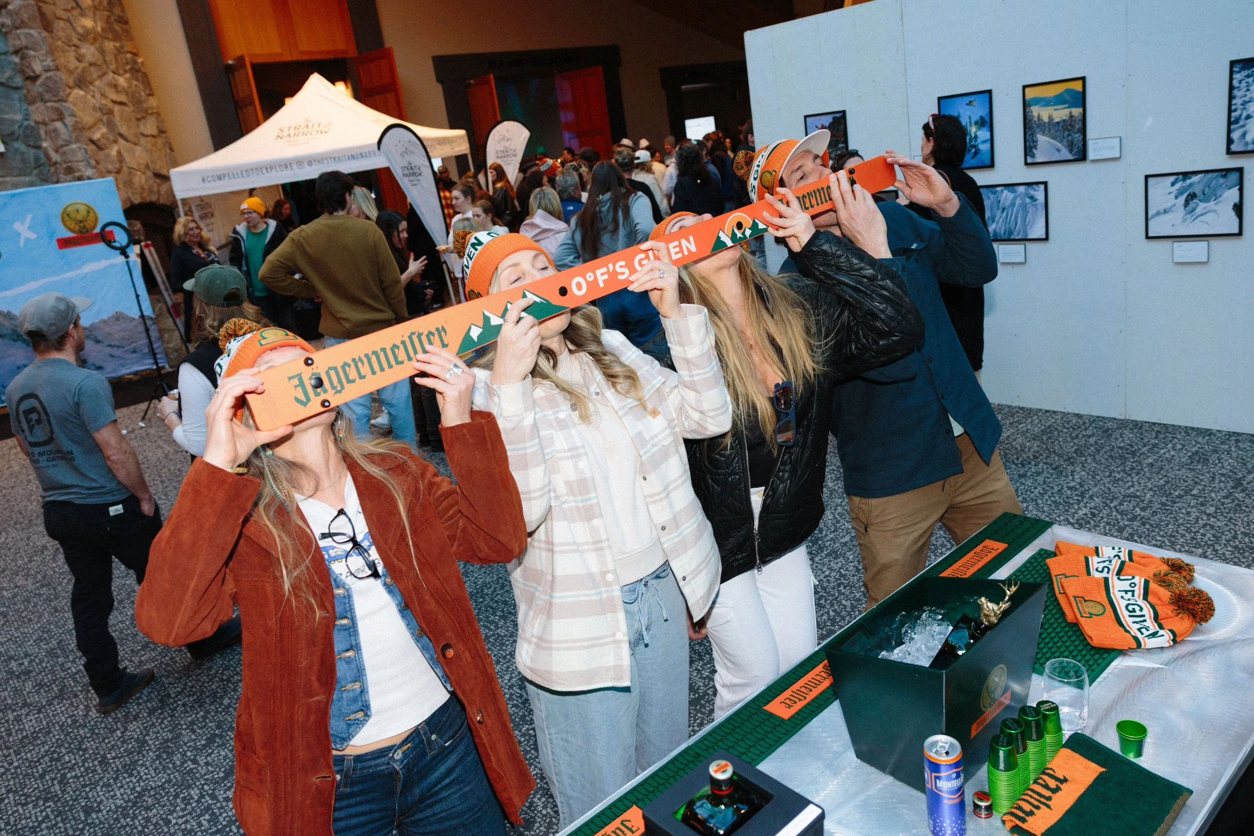 People at an event, with some women holding orange and green scarves that read 'Jägermüller Offs G' and 'Oefs G' in front of a table with merchandise, in a room with framed photographs on the wall.