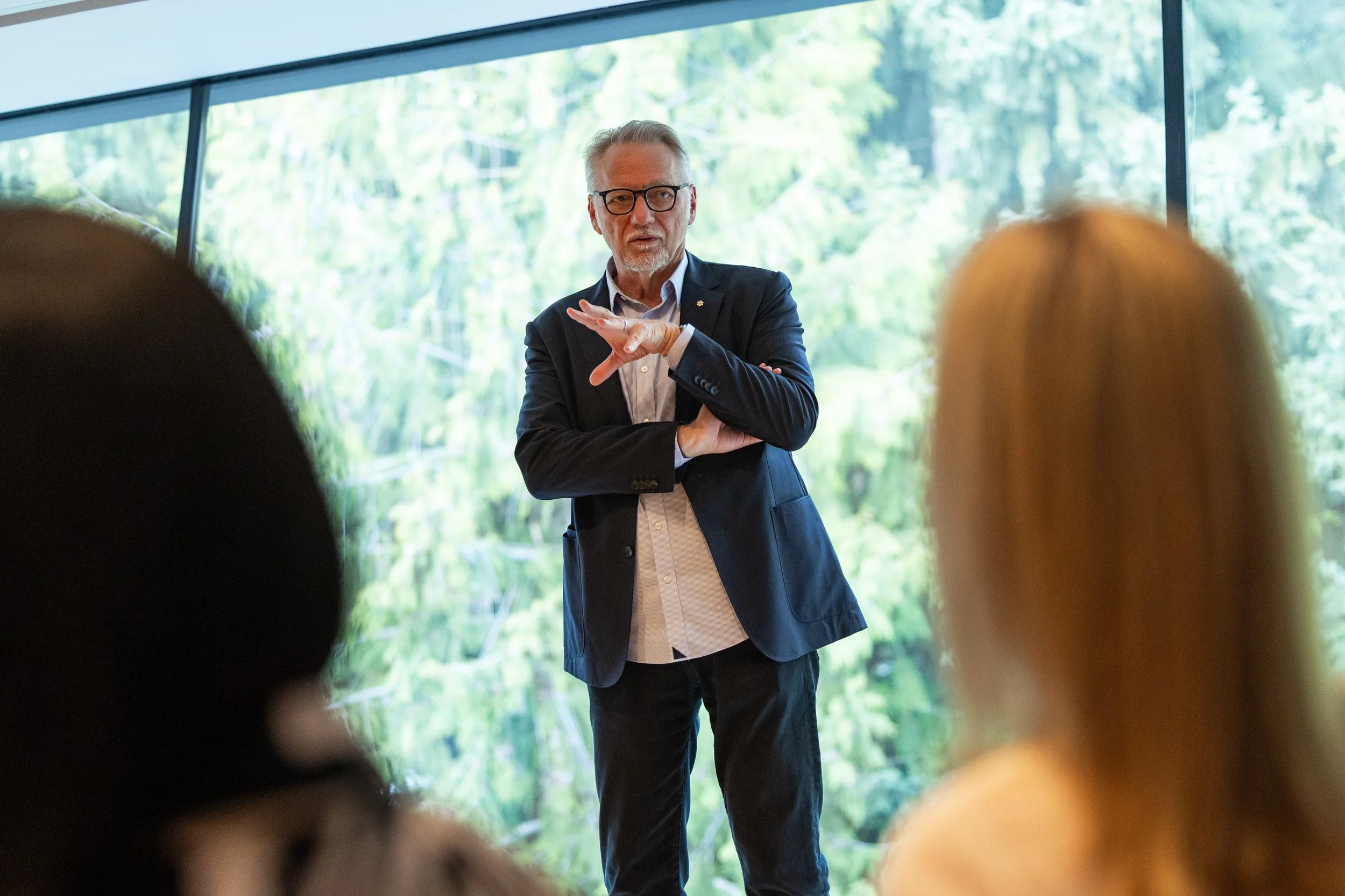 A man in a dark suit and glasses giving a presentation in front of large windows showing trees outside, with audience members in the foreground.