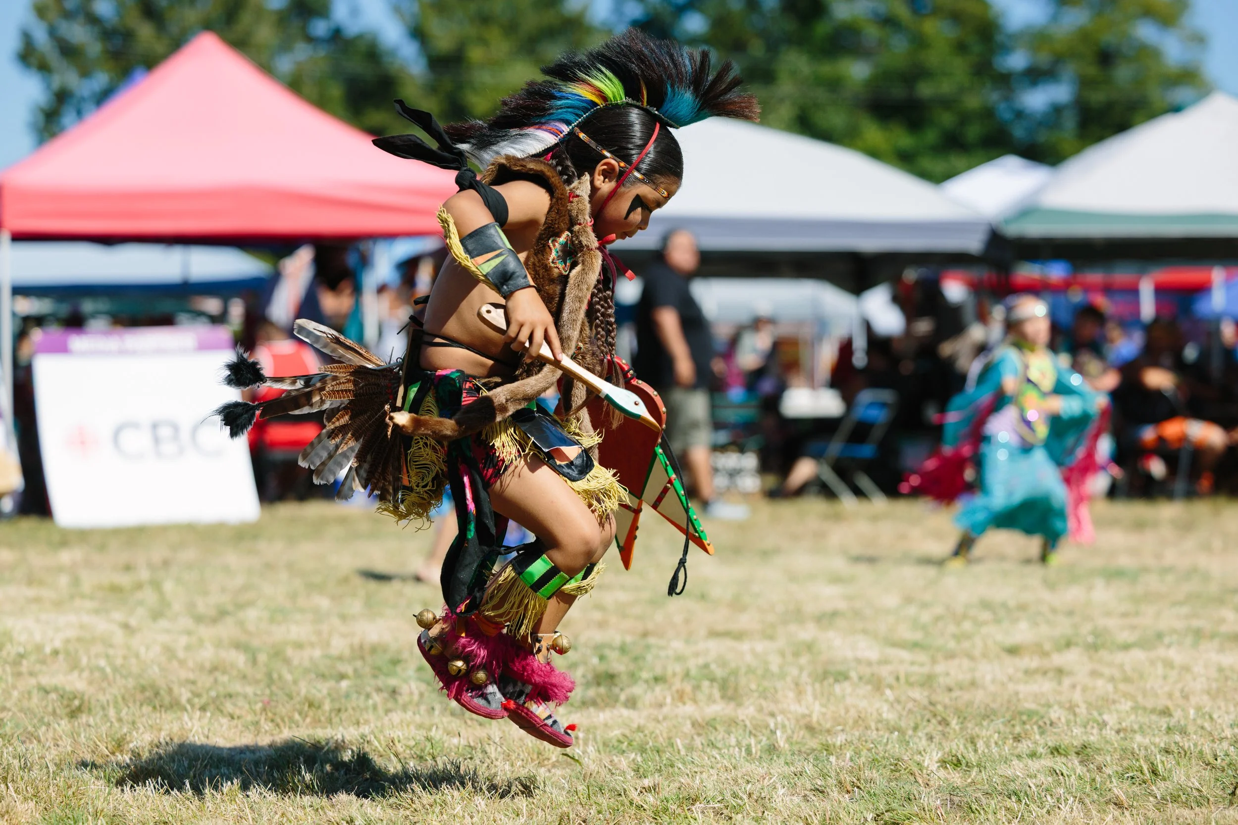 A young person wearing traditional Indigenous attire, including feathers and colorful accessories, jumping in the air during a cultural dance at an outdoor event with tents and spectators in the background.
