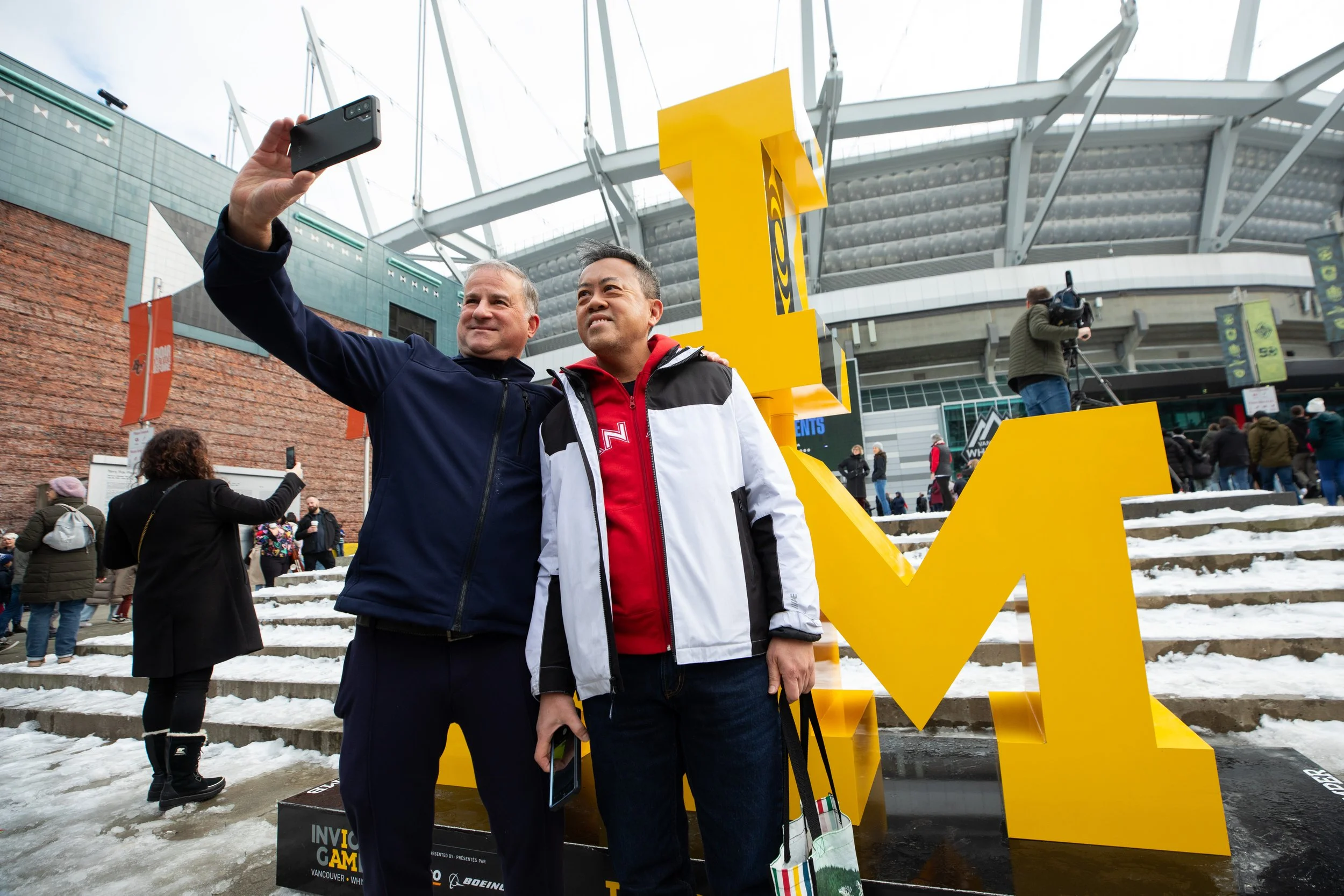 Two men taking a selfie in front of a large yellow down arrow at an outdoor event with snow on the steps and a crowd in the background.