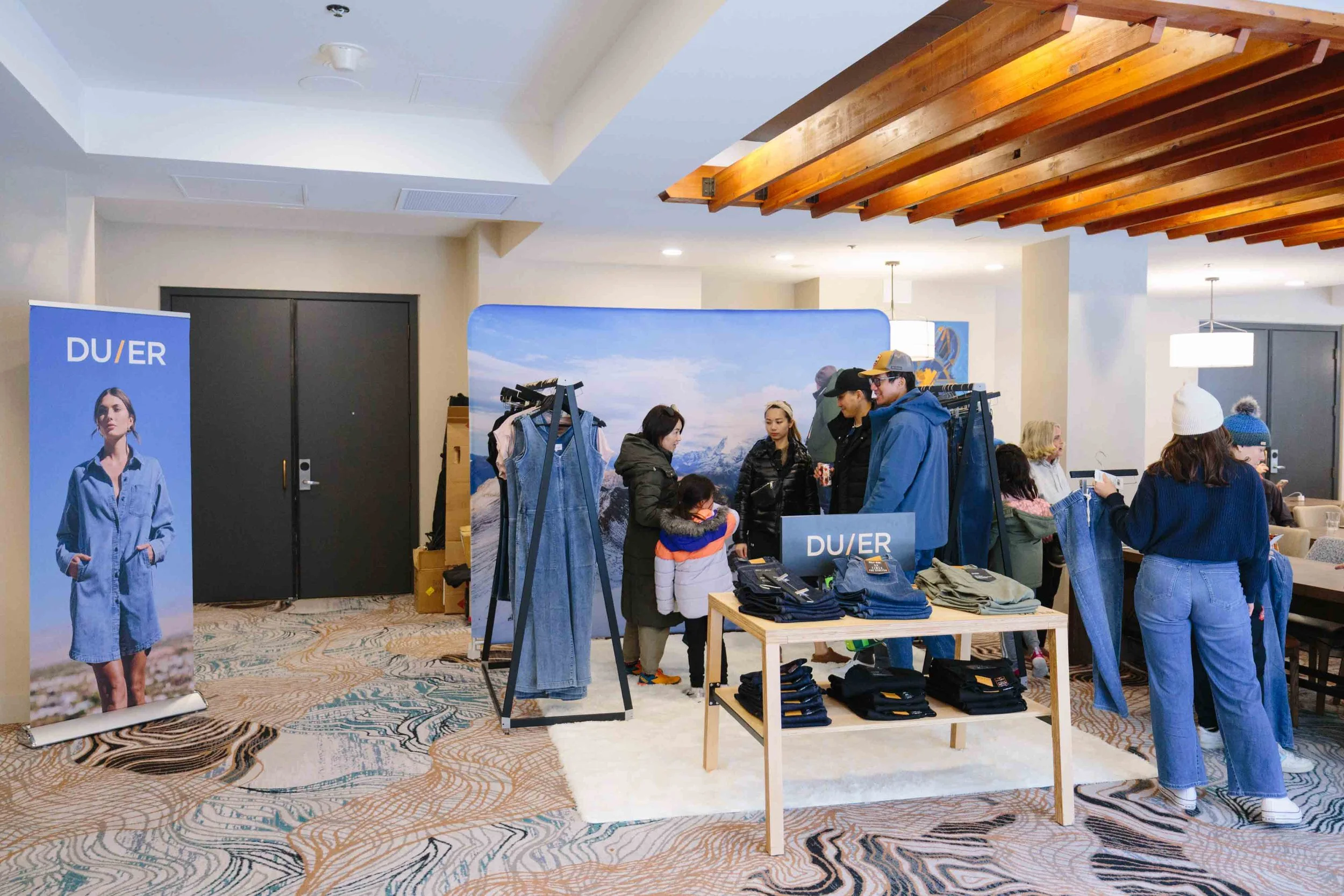 People shopping at a DU/ER clothing display in an indoor setting with a mountain landscape backdrop, tables with folded clothes, and promotional banners.