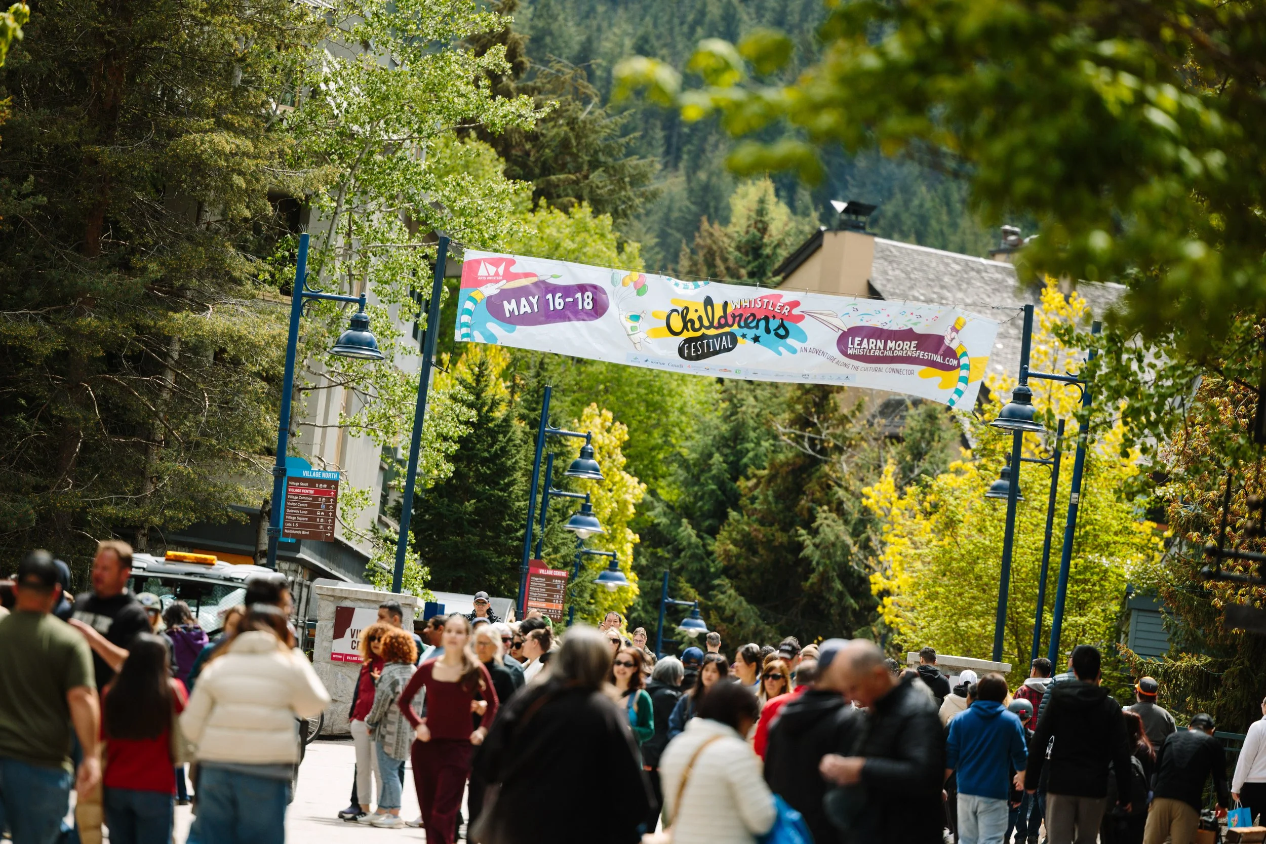 Crowd of people walking outdoors in a park during daytime, with trees and a festival banner overhead promoting the Whistler Children's Festival from May 16-18.
