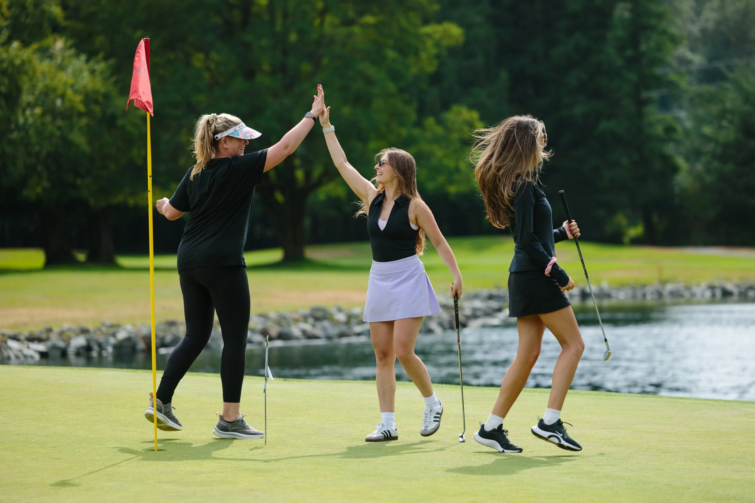 Three women on a golf course celebrating a successful game near a pond, one giving a high five to another, with a flagstick nearby and trees in the background.