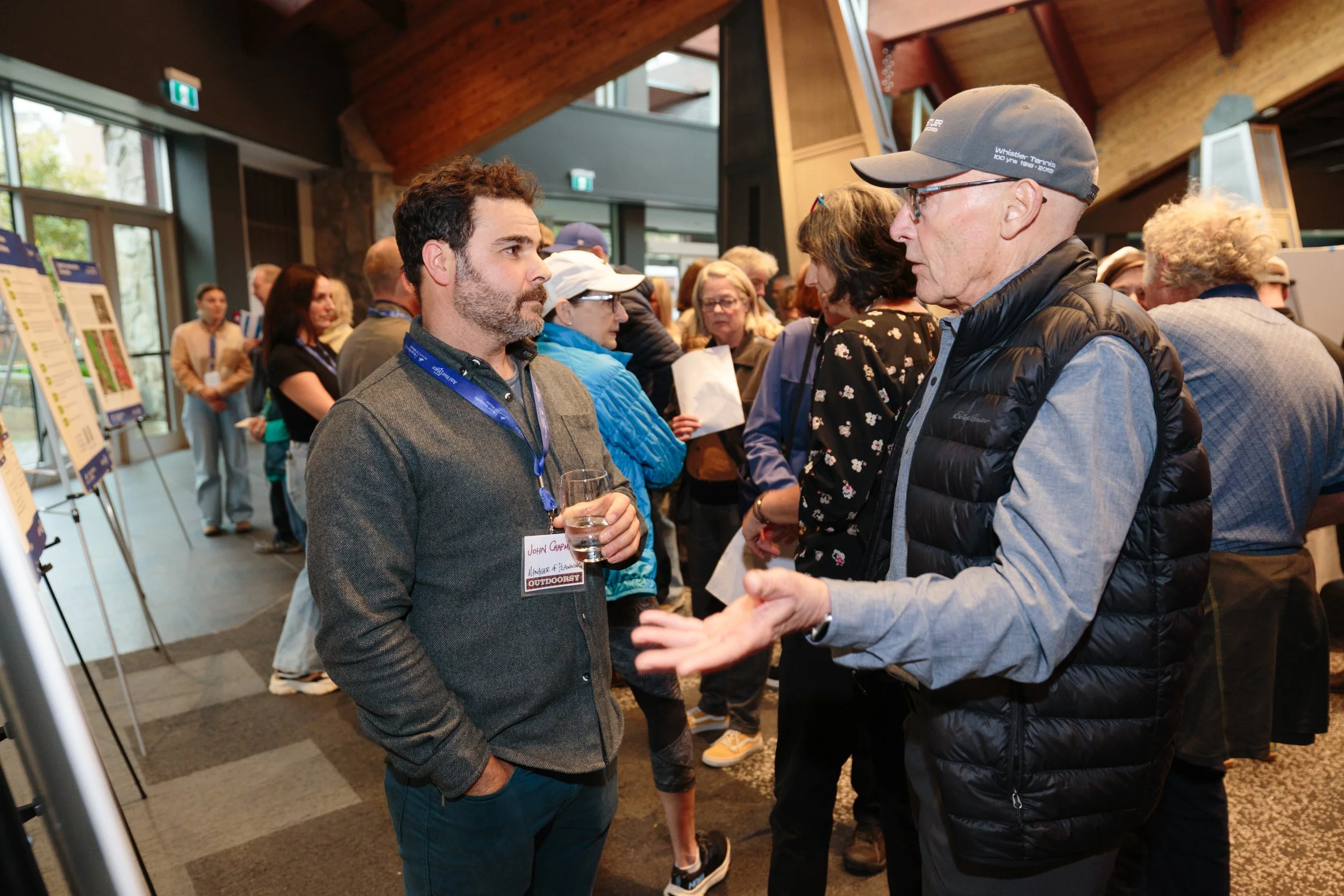 Two men are engaged in a conversation at a professional event, with other attendees and posters in the background.